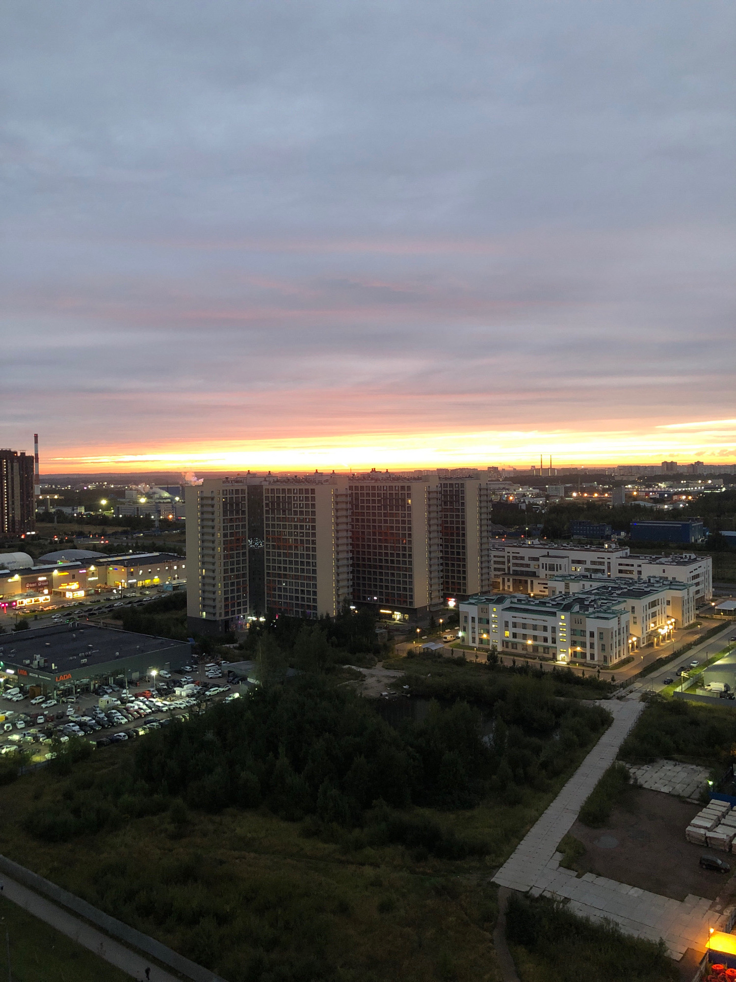 The image captures an aerial view of Novoye Devyatkino, Russia, at dusk. The sky is filled with a gradient of colors, transitioning from soft grays and purples at the top to a vibrant orange and yellow horizon. Several modern apartment buildings dominate the middle ground. One particularly large, rectangular building with many windows is centrally located, flanked by other similar structures. To the left, a taller, darker building with a prominent vertical structure rises against the sky. In the foreground and to the left, a large commercial building with illuminated signs is visible, adjacent to a busy parking lot filled with numerous cars. A dense area of trees and vegetation occupies the lower portion of the image, with a pathway or road winding through it. Several smaller buildings, some appearing to be industrial or commercial, are scattered throughout the cityscape in the background. The overall scene depicts a suburban or semi-urban environment during the transition from day to night.