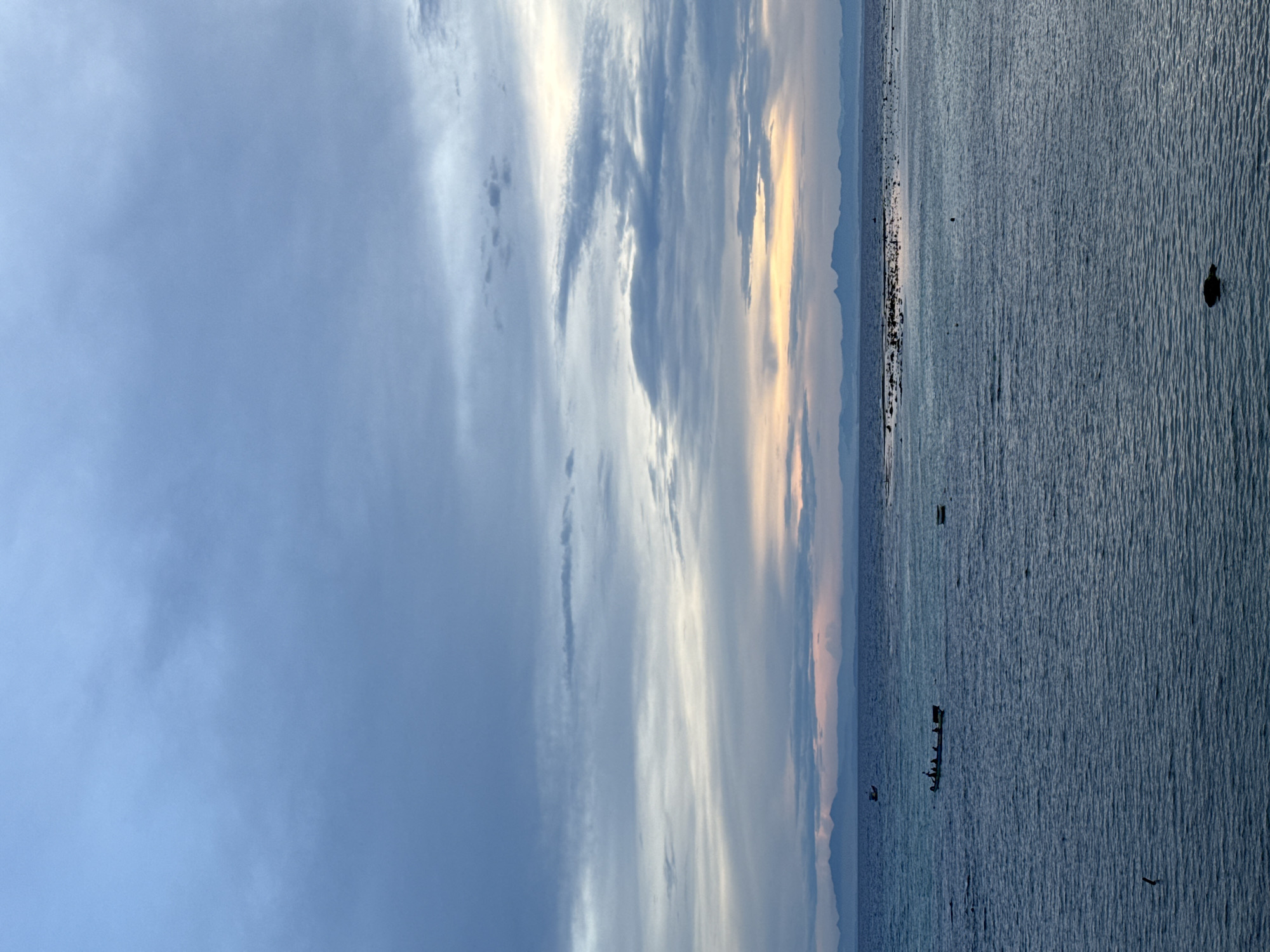 The image depicts a serene coastal scene at dusk, likely near the Lý Sơn Special Administrative Region in Vietnam. The sky is filled with a gradient of soft blues and grays, with hints of pale yellow and pink near the horizon, indicating the setting or rising sun. Wispy clouds stretch across the sky, adding texture to the atmospheric perspective.
The sea is a deep blue, with gentle ripples reflecting the muted light. In the distance, a silhouetted mountain range forms the backdrop. Several small boats are visible on the water. One boat, a long, narrow vessel, carries several people and is moving across the frame from left to right. Another, smaller boat is anchored further out. In the middle distance, a cluster of rocky outcrops or a reef is visible just breaking the surface of the water. The foreground shows a dark, indistinct object in the water, possibly a submerged rock or debris. The overall impression is one of tranquility and natural beauty.