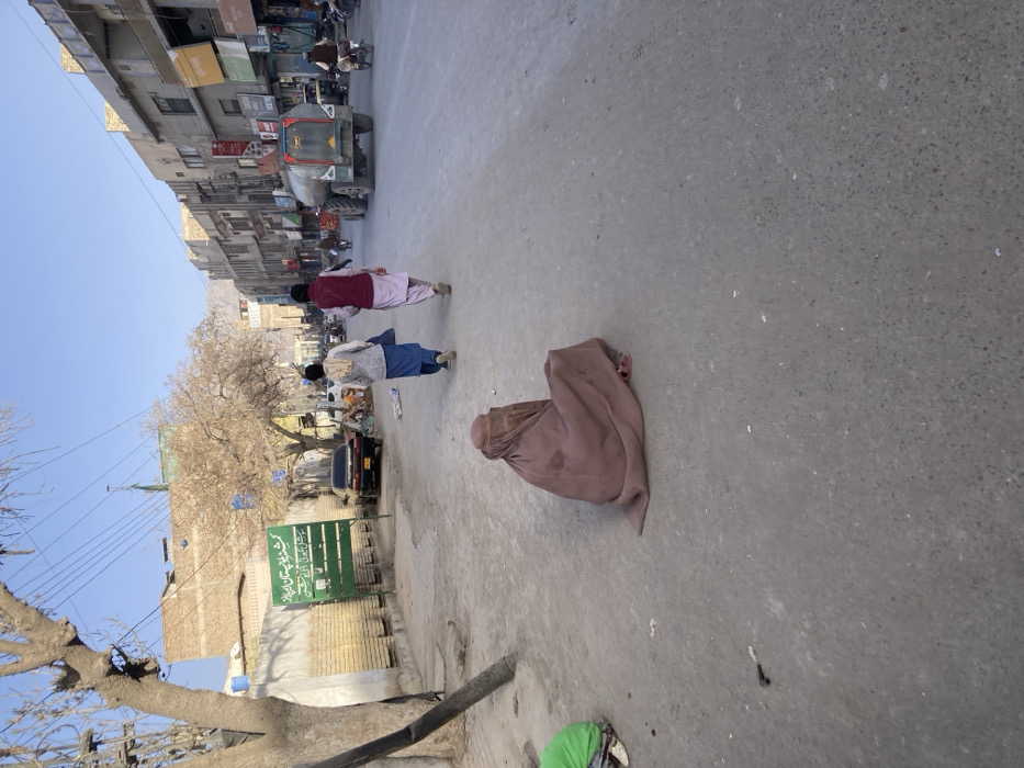 The image depicts a street scene in Quetta City Tehsil, Pakistan, under clear daylight. In the immediate foreground, a person in a green garment is partially visible, squatting on a dirt shoulder to the left. A woman, fully clad in a light brown burqa or niqab, is seated on the grey asphalt road, facing away from the camera and slightly to the right. Two males are walking away from the camera in the middle of the road; the individual on the left wears a light-colored top and blue trousers, while the individual on the right wears a maroon vest over a light shalwar kameez. To the left of the road, a green sign with white Urdu text is visible, behind which a dark sedan is parked. The right side of the road is lined with multi-story commercial buildings featuring various storefronts. Further down the street, a large water tanker truck and a motorcycle are observed moving away from the viewer. Overhead power lines are visible against a clear blue sky. Distant mountains frame the background.