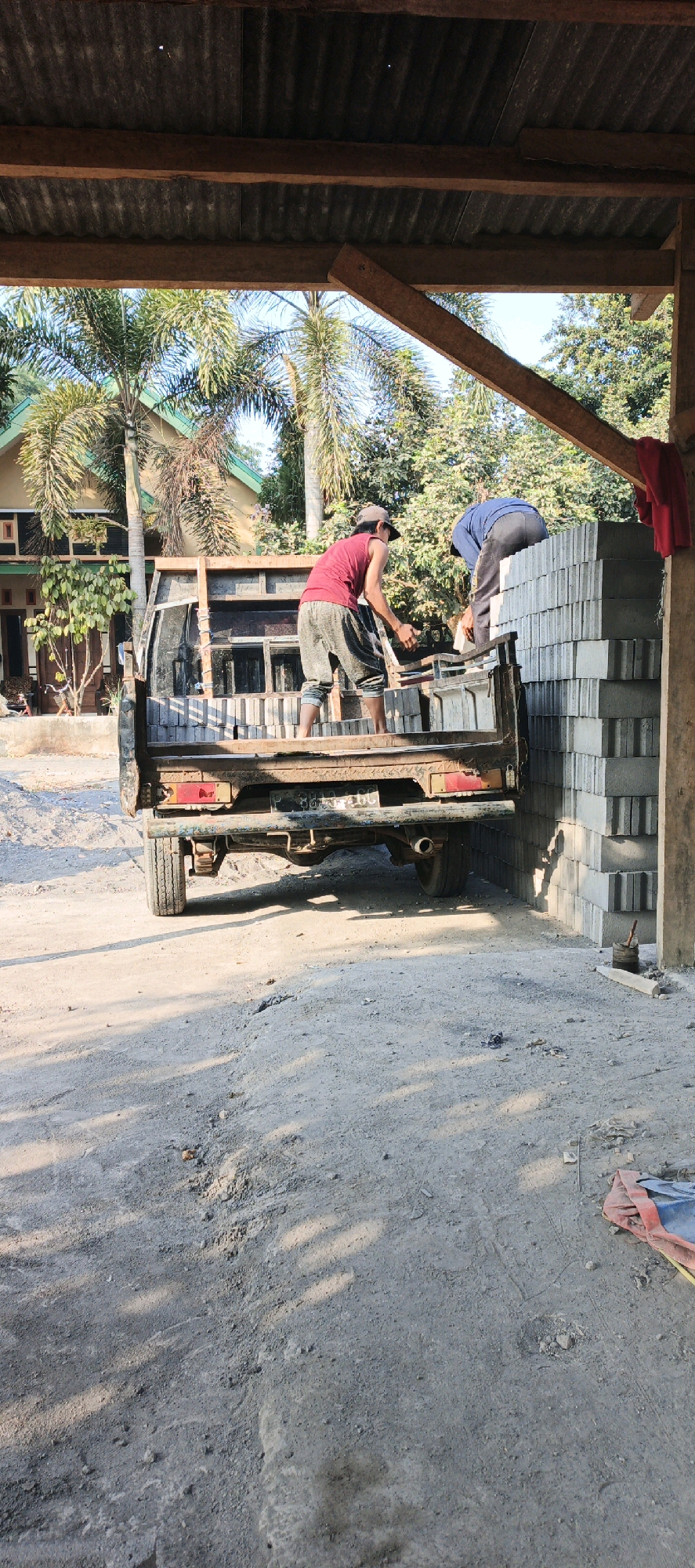 In Kasiyan, Indonesia, two individuals are engaged in unloading construction materials from the bed of a pickup truck. The truck is positioned under a wooden and corrugated metal shelter. One person, wearing a red tank top and grey pants, stands on the truck bed, actively handling rectangular blocks. The other, dressed in a blue long-sleeved shirt and grey pants, is positioned to the right of the truck, also interacting with the blocks. Stacked concrete blocks are visible to the right of the truck, forming a tall wall. The ground is dirt and gravel, with shadows cast by the shelter and trees. In the background, a house with a green roof and palm trees are present, indicating a residential or semi-rural setting. The overall scene depicts a moment of manual labor during a construction or renovation project.