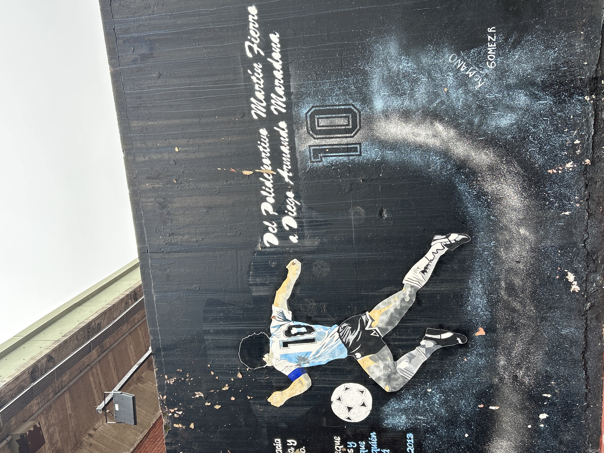 A mural of Argentine soccer player Diego Maradona is painted on a dark, rough-textured wall. Maradona is depicted in action, wearing his iconic number 10 jersey, dribbling a soccer ball. The mural is located in Buenos Aires, Argentina, on a wall beneath an overpass. The wall is weathered and shows signs of peeling paint and graffiti. The overpass, made of concrete and green metal, is visible in the background. The sky is overcast. Text in white paint above Maradona reads "Del Polideportivo Martin Fierro a Diego Armando Maradona". Below the mural, the names "ROMANO" and "GOMEZ.R" are also visible. The overall scene suggests a public art display in an urban environment.