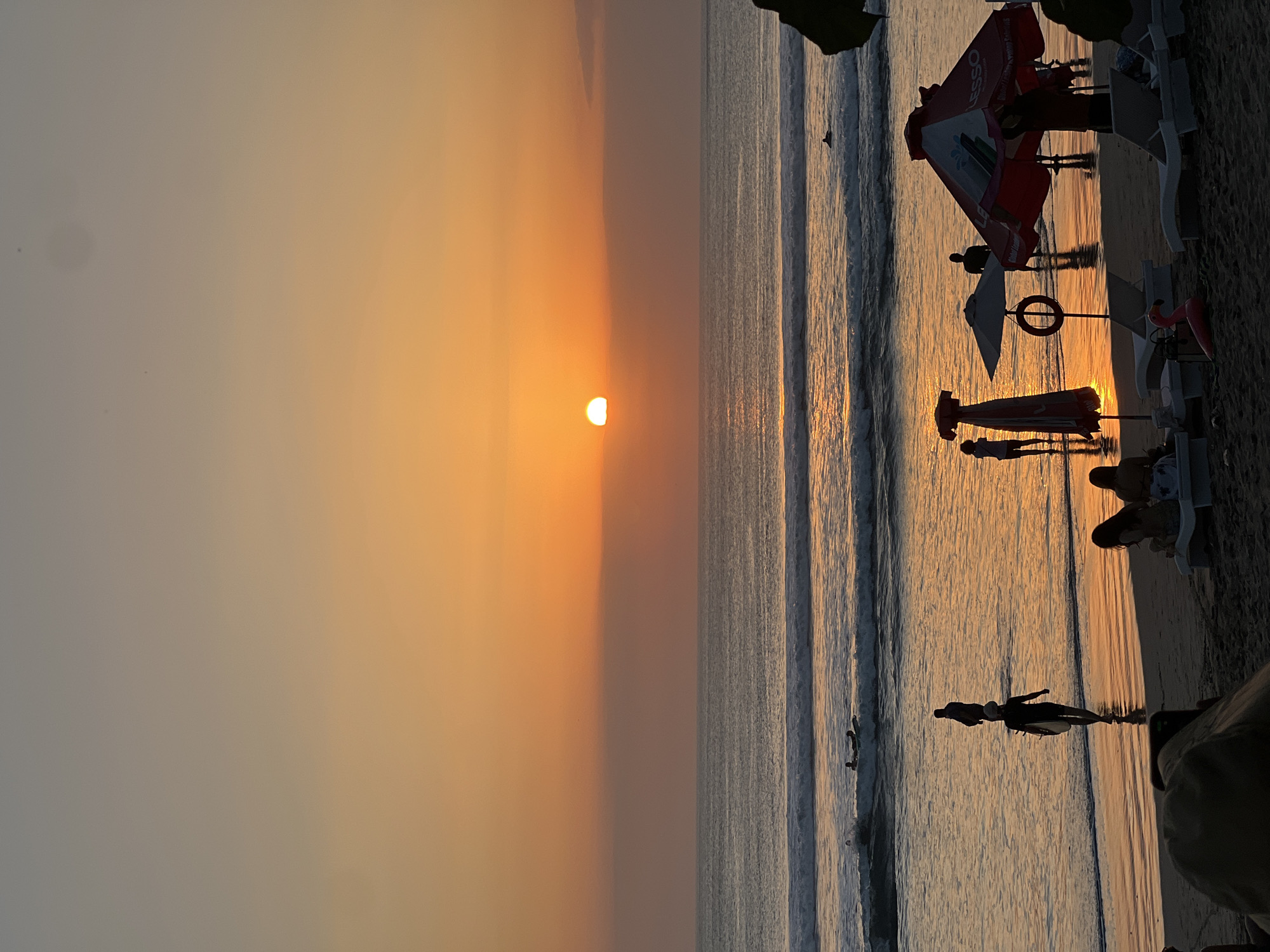 The image depicts a sunset on Kuta Beach, Bali, Indonesia. Silhouetted figures walk along the wet sand near the water's edge, some carrying surfboards. Several beach umbrellas and lounge chairs are visible on the drier sand in the foreground. The sun, a bright orange orb, is setting over the ocean, casting a warm glow on the water and the sky. Waves are breaking on the shore, and a few surfers are in the water. The overall atmosphere is peaceful and serene, characteristic of a tropical beach at dusk.