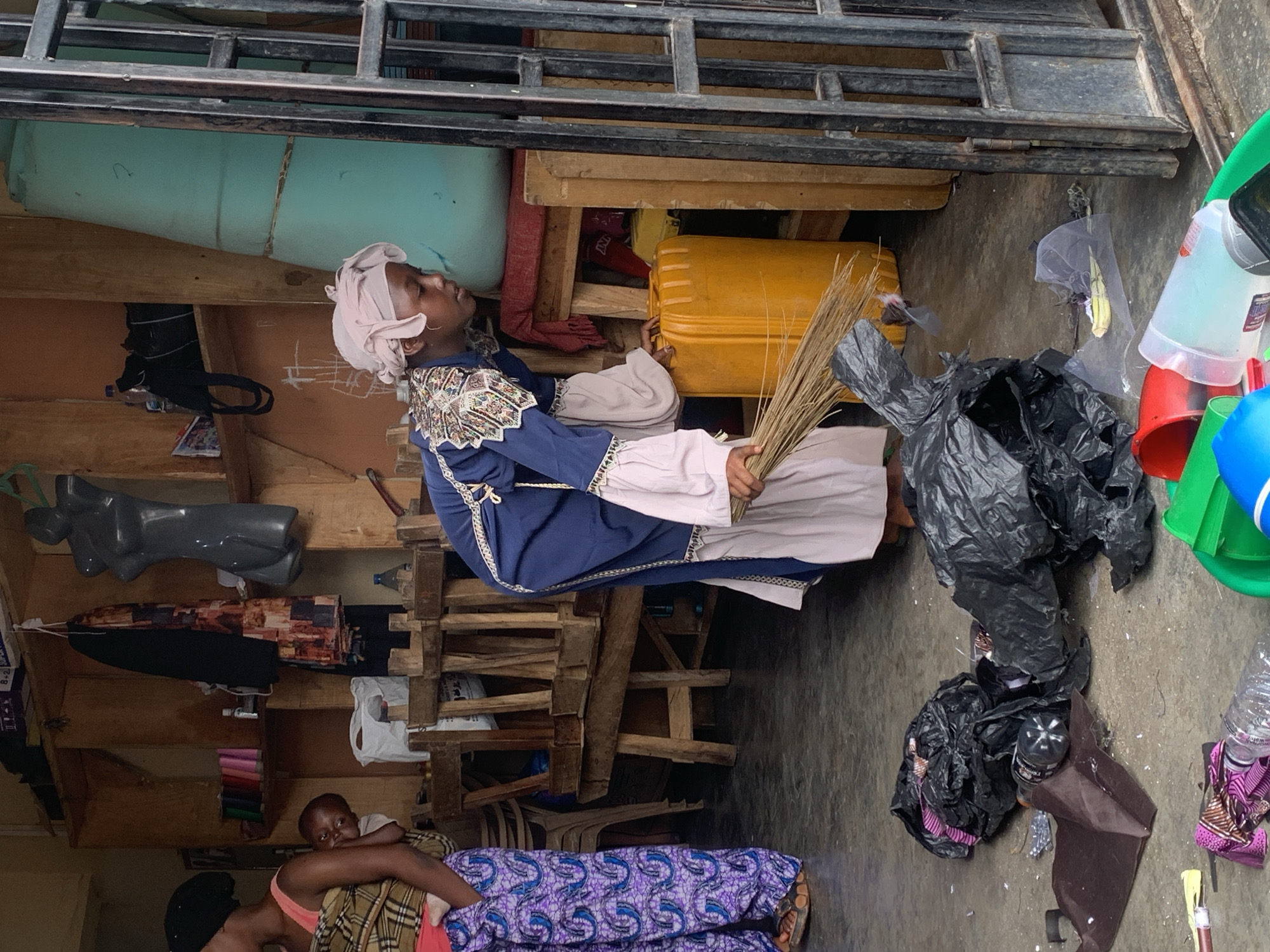 The image depicts an indoor scene, possibly a market stall or workshop, in Jos, Nigeria. The lighting suggests daytime. In the foreground, two people are visible. One person, wearing a pink top and a patterned wrap, is lying down and appears to be holding a baby or a small child. The child is looking towards the camera with a neutral expression.
Behind them, a young person wearing a blue robe with white trim and a pink headscarf is holding a broom made of dry grass. This person is looking upwards and to the right, their expression is unreadable. The background is cluttered with various objects, including shelves filled with items, a large yellow plastic container, a large blue object that might be a water tank or drum, and several black plastic bags filled with what appears to be trash. A mannequin torso is also visible on a shelf. The overall setting appears somewhat disorganized, with items piled up on shelves and on the floor. The floor itself is concrete, and there are some scattered debris. The metal bars across the top of the image suggest it might be an open-fronted structure, such as a stall.