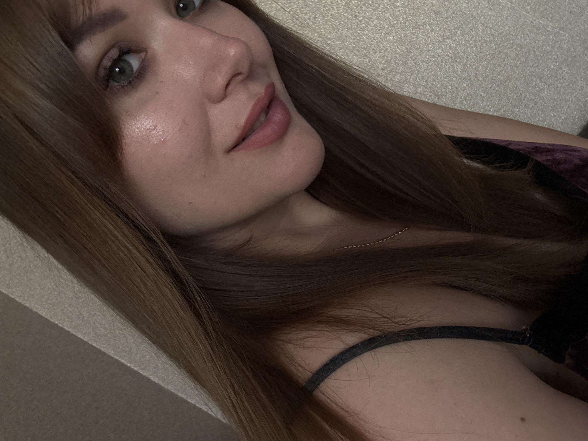 A close-up photograph shows a woman with light skin, light eyes, and long brown hair. Her face is turned slightly towards the camera, with a slight smile on her lips. She appears to be wearing light makeup. A thin necklace is visible around her neck, and a dark-colored tank top is partially visible on her shoulder. The background is a neutral, textured wall. The location in Chaikovsky, Russia, cannot be determined from the visual information.
