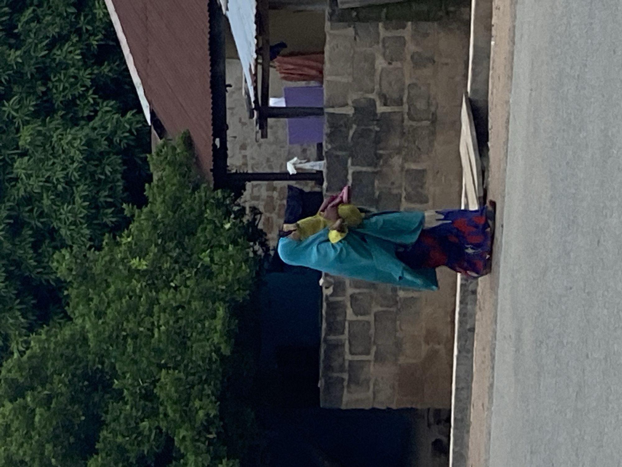 In Mararaba, Nigeria, a woman is carrying a load on her head. She is wearing a blue head covering and gown, and a red patterned dress. She is walking on a grey paved surface, adjacent to a building with stone-like construction. Green trees and foliage are on the left. A portion of a building with a corrugated roof is also visible.