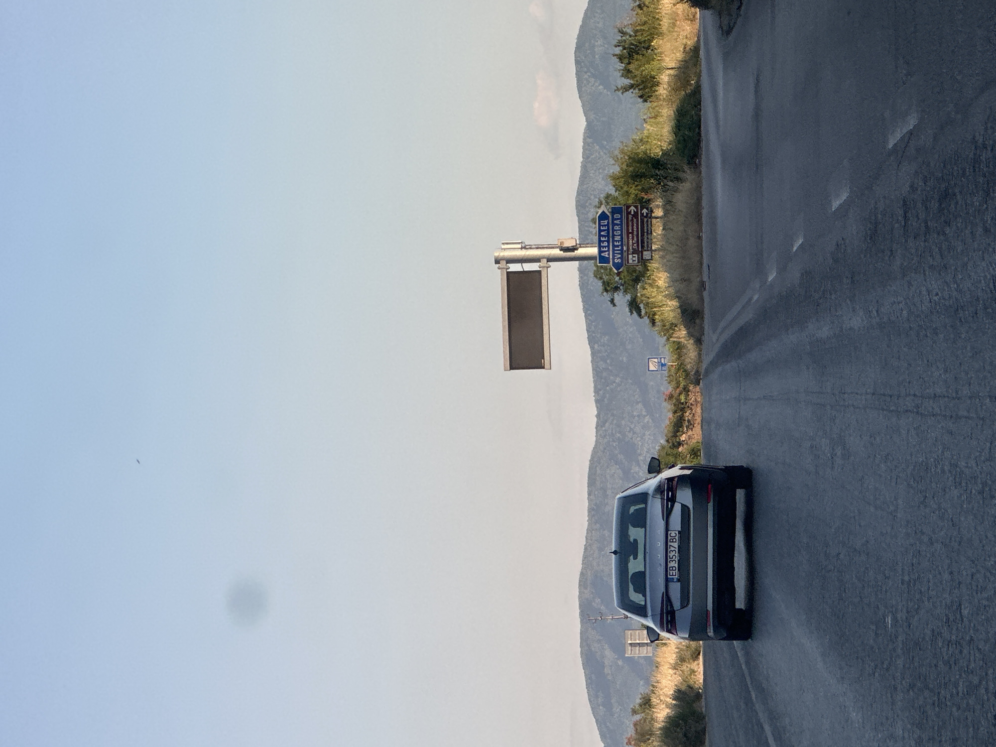 The image depicts a road scene with a car driving away from the viewer. The car is a silver sedan with a license plate reading "EB 3537 BC". The road is asphalt with faded white lane markings. On the right side of the road, there is a blue directional sign pointing towards "DEBELSKOV" and "SVILENGRAF". Further along the road, there is an electronic display board mounted on a pole. The background consists of rolling hills covered in vegetation and trees, with a clear blue sky above. A small dark speck, possibly a bird or insect, is visible in the sky on the left side of the frame. The overall impression is a rural or semi-rural environment in Bulgaria, possibly near the city of Prisovo, given the context of the directional sign. There are no people visible in the image, and the action is limited to the car driving on the road. The lighting suggests it is daytime, likely late afternoon due to the warm tones and long shadows.