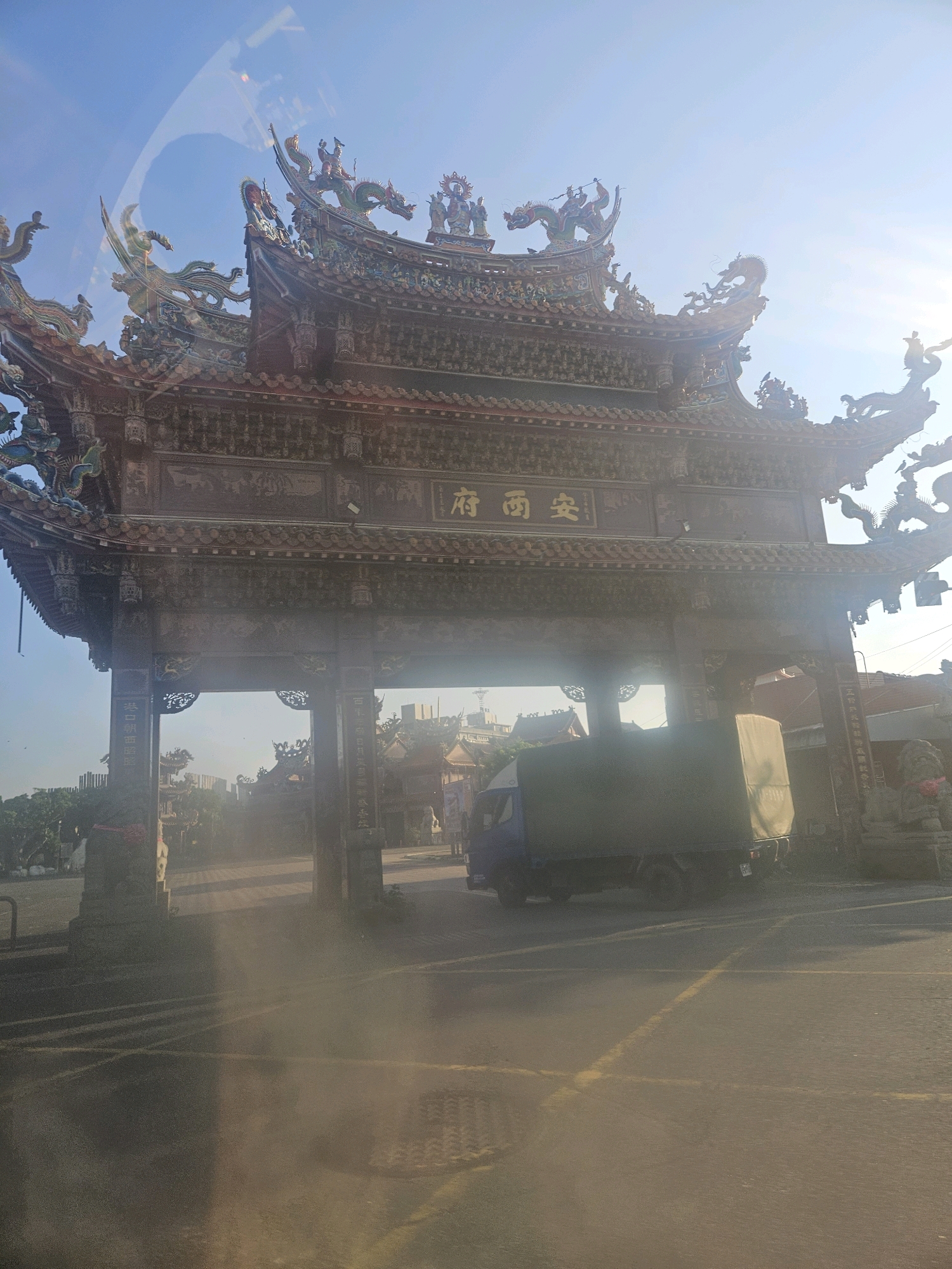 The image captures a large, ornate temple gate in Taixi Township, Taiwan, under a bright, clear sky. The gate, constructed of dark wood and adorned with intricate carvings and vibrant dragon figures, stands prominently in the foreground. Two stone lion statues flank the entrance. Through the archway of the gate, a blue truck with a canvas-covered cargo bed is visible, parked on what appears to be a paved road. The road is marked with yellow lines, and in the distance, other traditional Taiwanese buildings and trees can be seen. The overall scene suggests a sunny day in a culturally rich area.