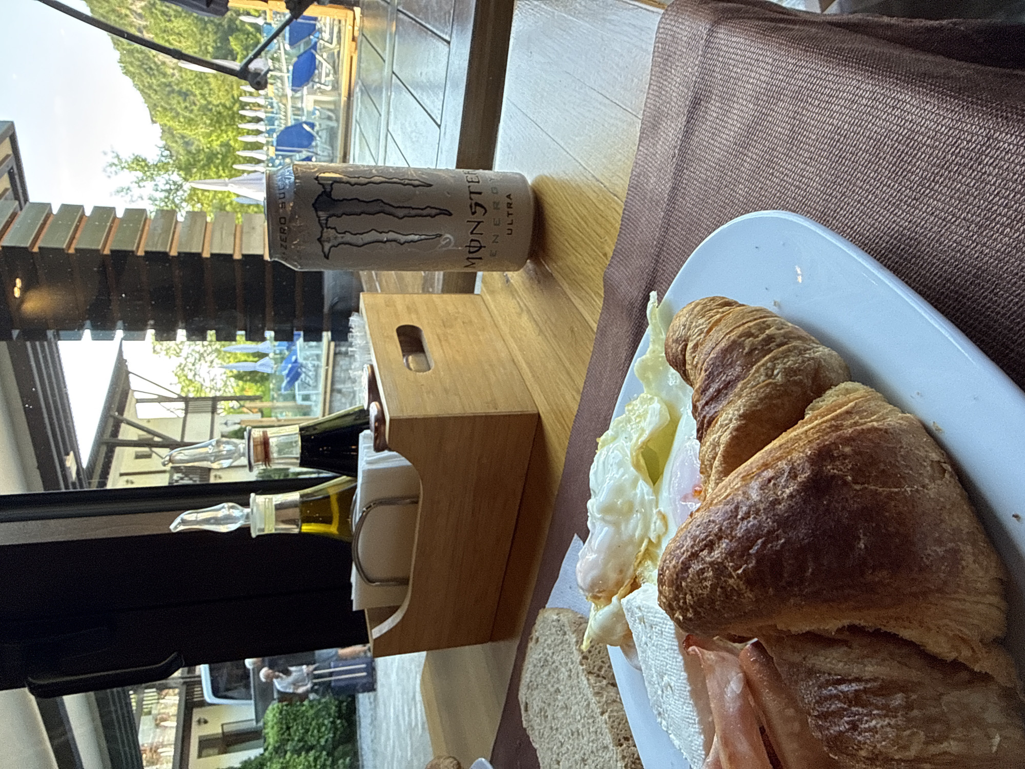 The image shows a table setting at a restaurant, likely in the city of Veliko Tarnovo, Bulgaria, based on the visible buildings and landscape in the background. The table is covered with a brown cloth and has a white plate containing a croissant sandwich with fried egg and ham. A can of Monster Energy Ultra drink rests near the plate. There is a wooden napkin holder and a set of oil and vinegar bottles. Through the window, a scene of a resort-like area with swimming pools and greenery can be observed. A person is visible through the glass window. The lighting is bright, suggesting a daytime shot.