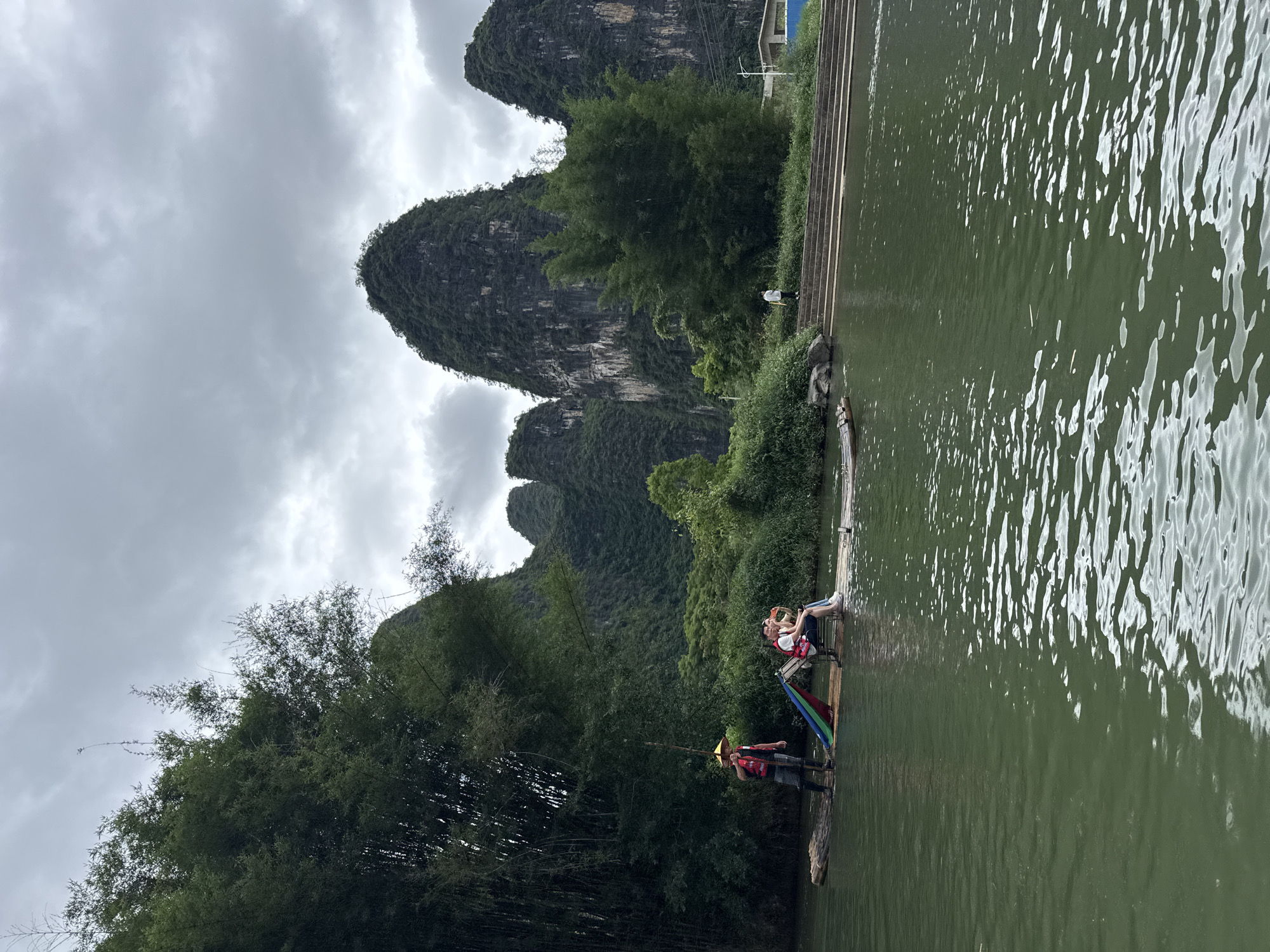 A landscape photograph reveals the natural beauty of Guilin, China. In the foreground, a bamboo raft floats on a river. Two tourists, dressed in white shirts and red life vests, sit on chairs on the raft, while a person wearing a straw hat stands at the back, likely navigating the raft. The river reflects the overcast sky, causing a slight sheen. Towering karst mountains covered in green vegetation are visible in the background, adding to the scenic charm. Lush green trees flank the riverbanks on both sides. A set of stairs are visible on the right side of the photograph that lead down to the riverbank.