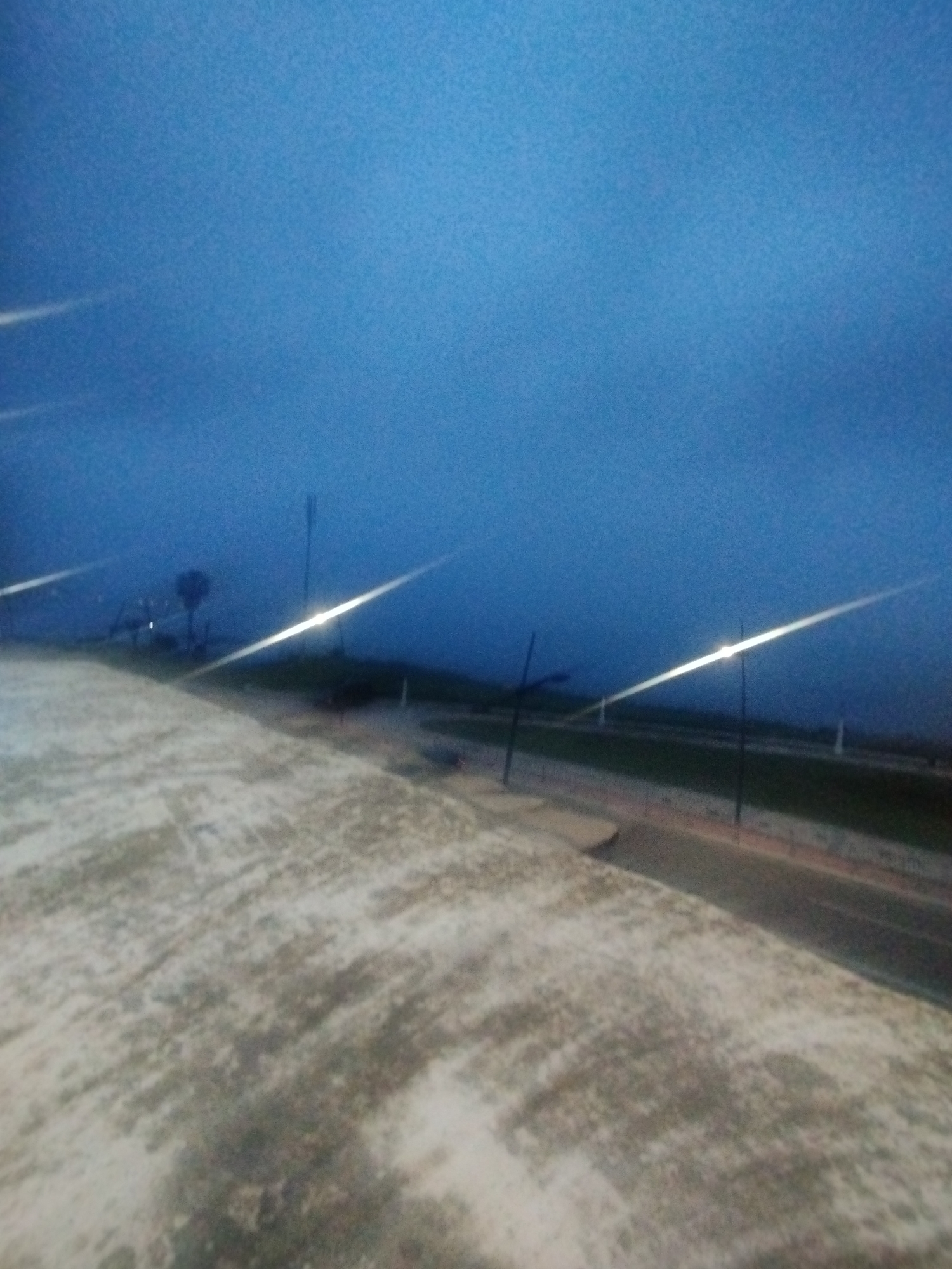 This media file presents an outdoor scene captured during twilight or nighttime in Fnideq, Morocco, from an elevated viewpoint, possibly a rooftop or balcony. The foreground features a rough-textured, light-colored surface, resembling concrete or a paved area, displaying patches of various gray and brown hues.
In the mid-ground, a dark strip, likely a road or path, runs horizontally. Several artificial light sources, possibly streetlights, are prominent, creating intense, elongated streaks of bright white light that cut across the frame, suggesting a long exposure or lens flare. Slender poles are also visible in proximity to these lights.
The background shows a distant landscape or horizon largely obscured by haze or fog, appearing as a dark, indistinct mass against the sky. A lone tree and a faint, dark structure can be discerned on the left side of this hazy horizon. The sky occupies the upper portion of the image, displaying a deep, uniform blue color characteristic of dusk or dawn. No people are visible, and there is no discernible text. The overall impression is one of tranquility and dim visibility, typical of a coastal area at night or twilight.