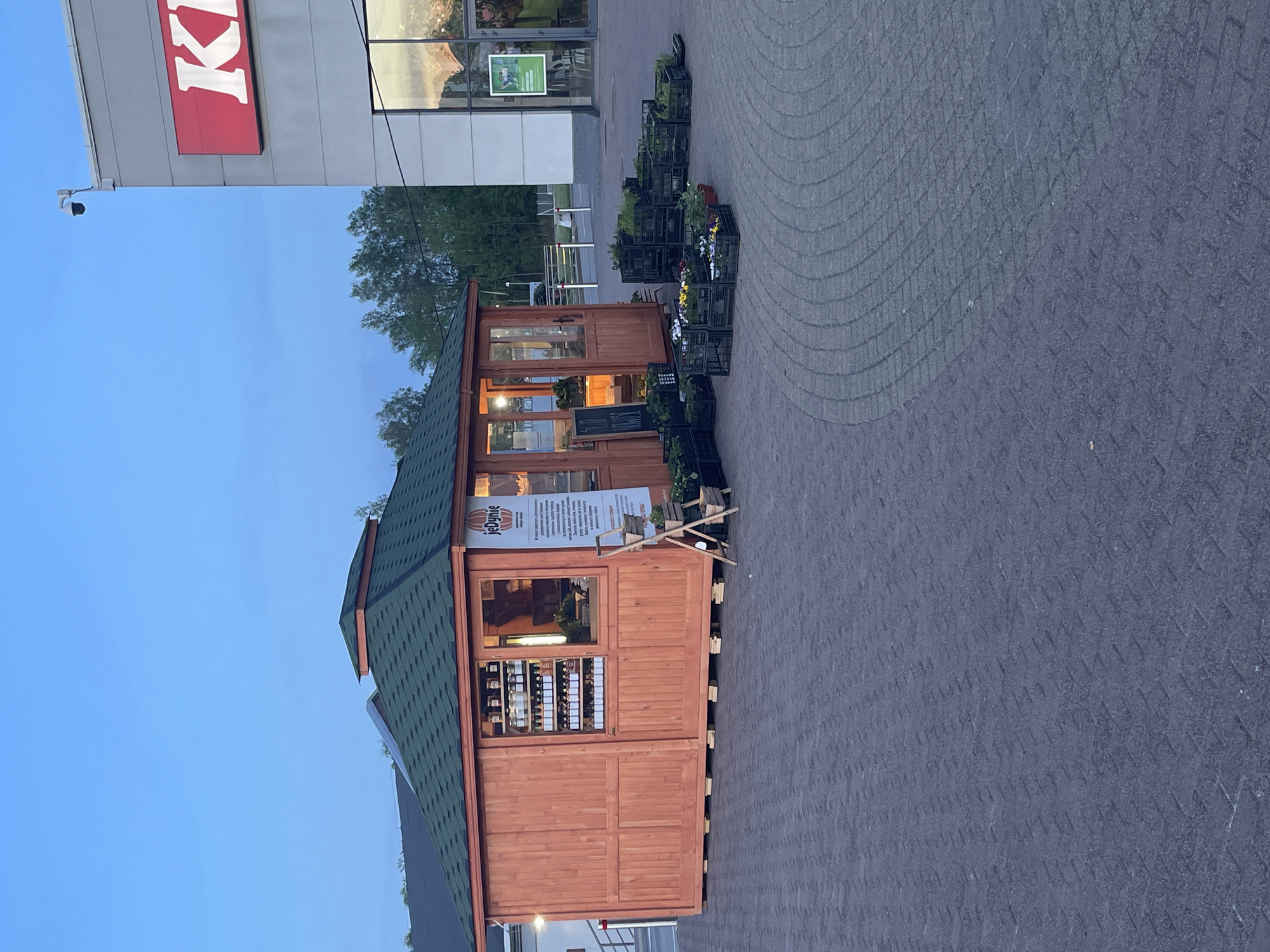 The image shows a small wooden building, likely a retail space, with a green shingle roof and glass windows displaying merchandise. The building is situated on a paved surface with a pattern suggestive of interlocking pavers. Adjacent to the building, black crates containing plants are arranged. In the background, there's a larger building with a sign that partially reads "KI," possibly indicating a store like KFC. The sky is visible in the background, suggesting an outdoor setting. The location is described as being in the city of Unknown, Unknown.