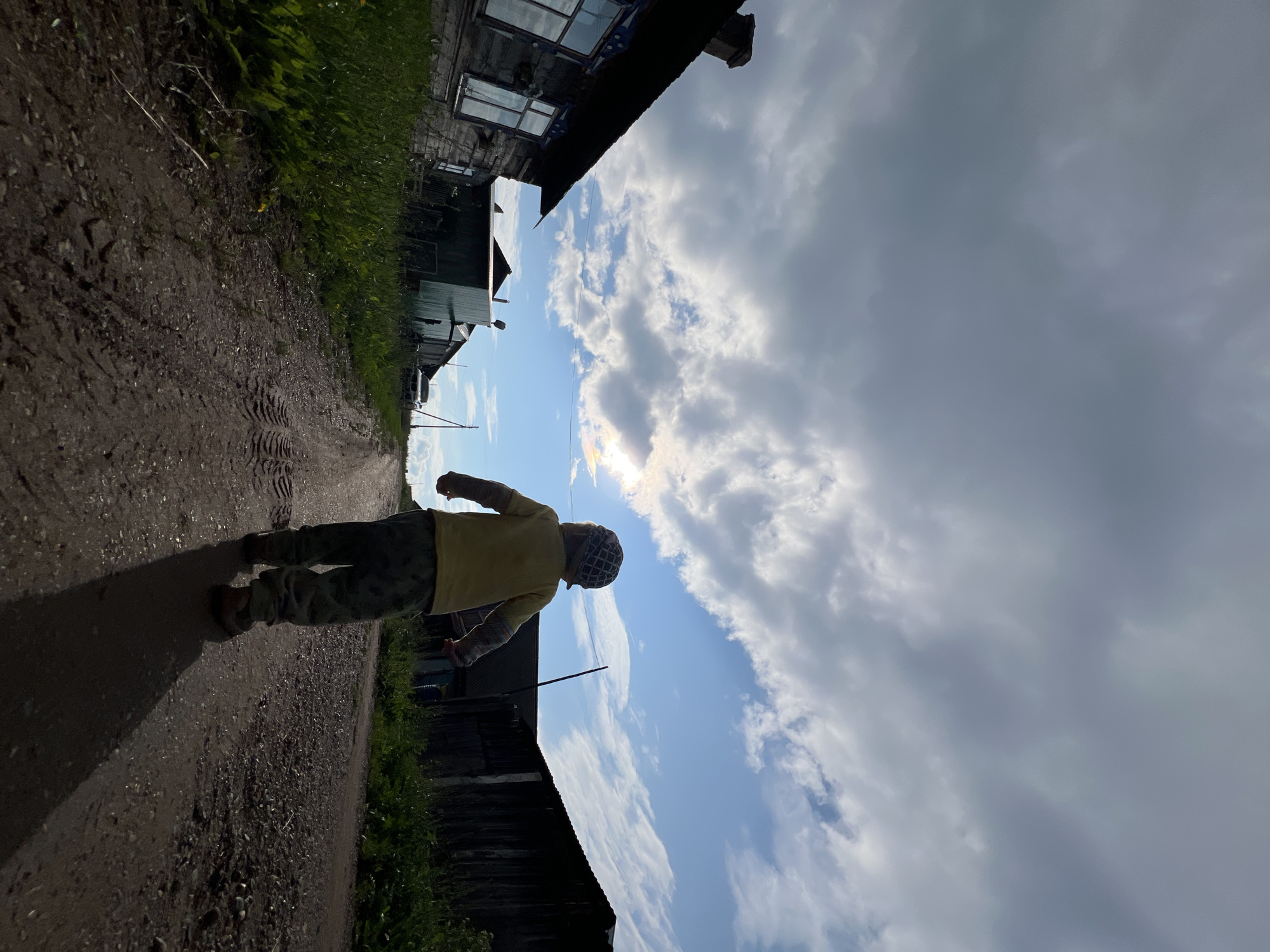 In Panteleevka, Russia, a low-angle shot captures a young child walking away from the camera down a dirt road. The child, wearing a yellow shirt and patterned pants and hat, casts a long shadow on the road. To the left, a weathered wooden house with ornate window trim stands, its windows reflecting the sky. To the right, a dark wooden structure, possibly another house or shed, is visible. Tall green grass lines the sides of the road, and in the distance, a white vehicle is parked. The sky is dramatic, with a mix of heavy grey clouds and patches of bright blue, illuminated by the sun which creates a faint rainbow halo effect. The overall scene suggests a rural setting in Panteleevka under a dynamic sky.