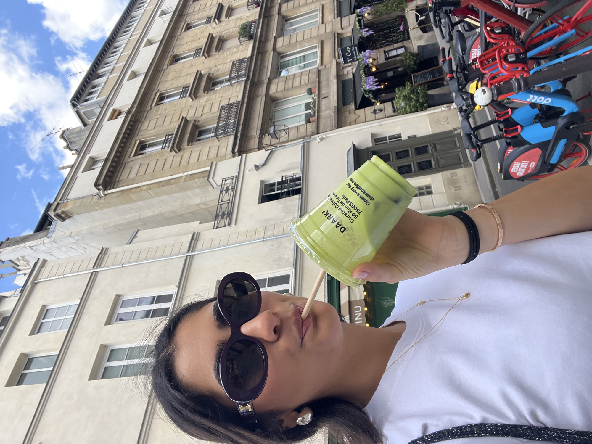 A woman wearing sunglasses is drinking from a plastic cup with a straw. The text on the cup reads "DAARK3 Curated Coffee Con 50 Rue de Turenne 75003 Paris Open every day daarkcoffee.com." The woman is wearing a white t-shirt, a gold bracelet, a black hair tie, and a gold necklace. The background shows a building with windows and bikes. The bikes are branded with the word "DOTT" and the phrase "ESSAI GRATUIT!" The building appears to be located on Rue de Turenne in Paris, France.