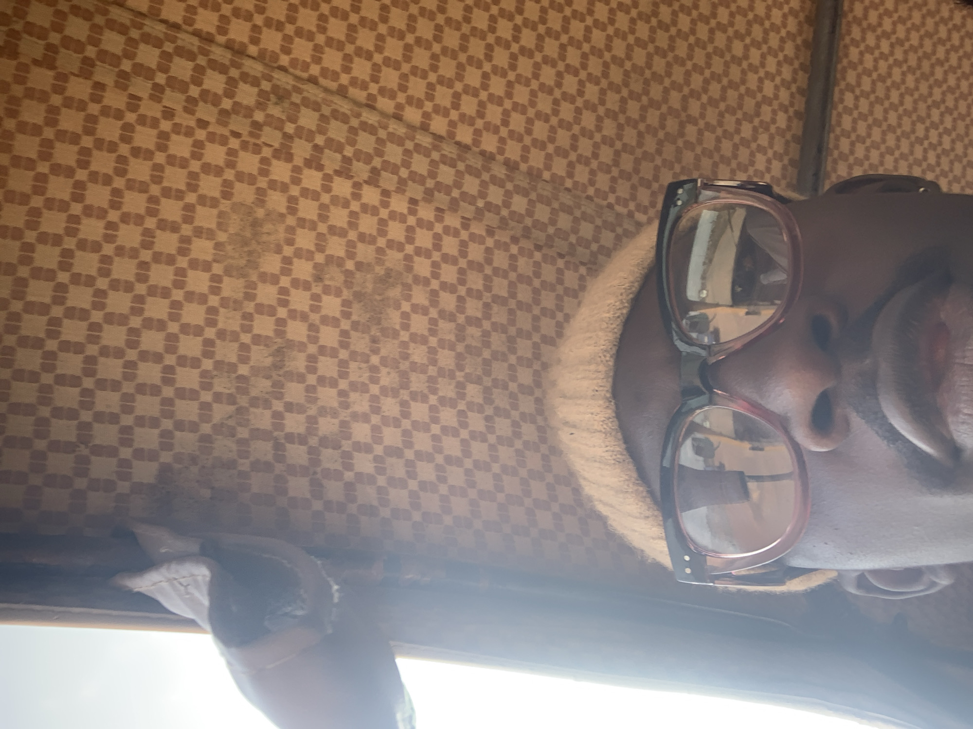 A close-up, low-angle, indoor shot shows the face of a Black individual from the nose upwards. They are wearing a light-colored beanie and thick-rimmed glasses with pink frames. The background is a textured, checkered fabric in shades of beige and brown, suggestive of a vehicle's interior. A faint light source is visible on the left side, possibly a window. The scene is set in Nabatum, Nigeria.