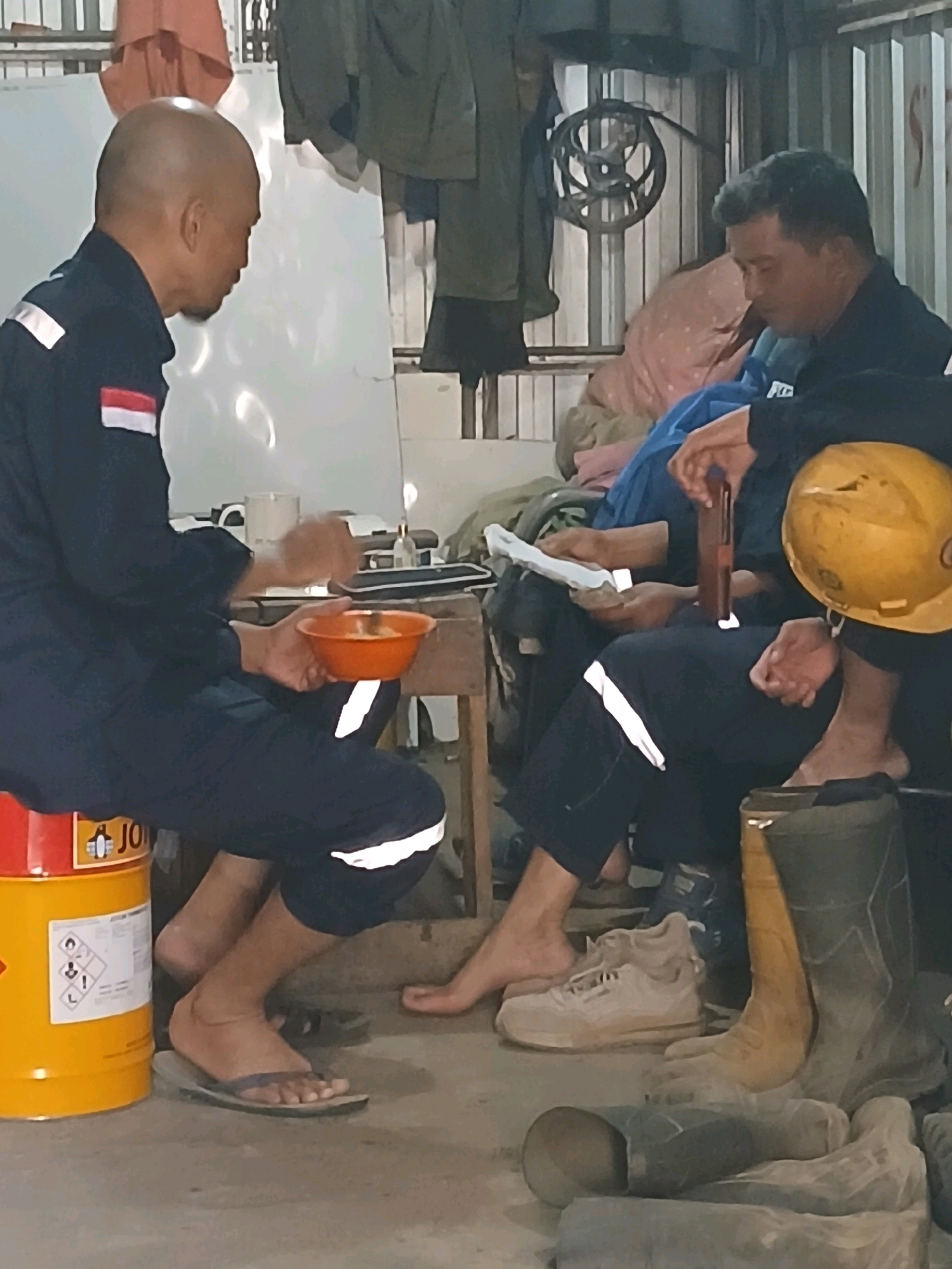 The image depicts a dimly lit indoor setting, possibly a workshop or break room, with a concrete floor. Three individuals are present, two of whom are seated. The person on the left, a bald man wearing a dark blue uniform with red and white stripes on the arm, is seated on a large yellow cylindrical container. He is holding an orange bowl and appears to be eating. To his right, another man in a similar dark blue uniform is seated, holding a piece of paper and a brown object. He has a yellow hard hat resting on his lap. A third person is partially visible in the background, seated and covered by a blue and pink fabric. Several pairs of boots and a pair of sneakers are on the floor in the foreground. The environment suggests a practical, possibly industrial or construction-related location. The presence of a hard hat and work boots points to a work environment. The overall impression is one of a brief respite or break during work hours.
