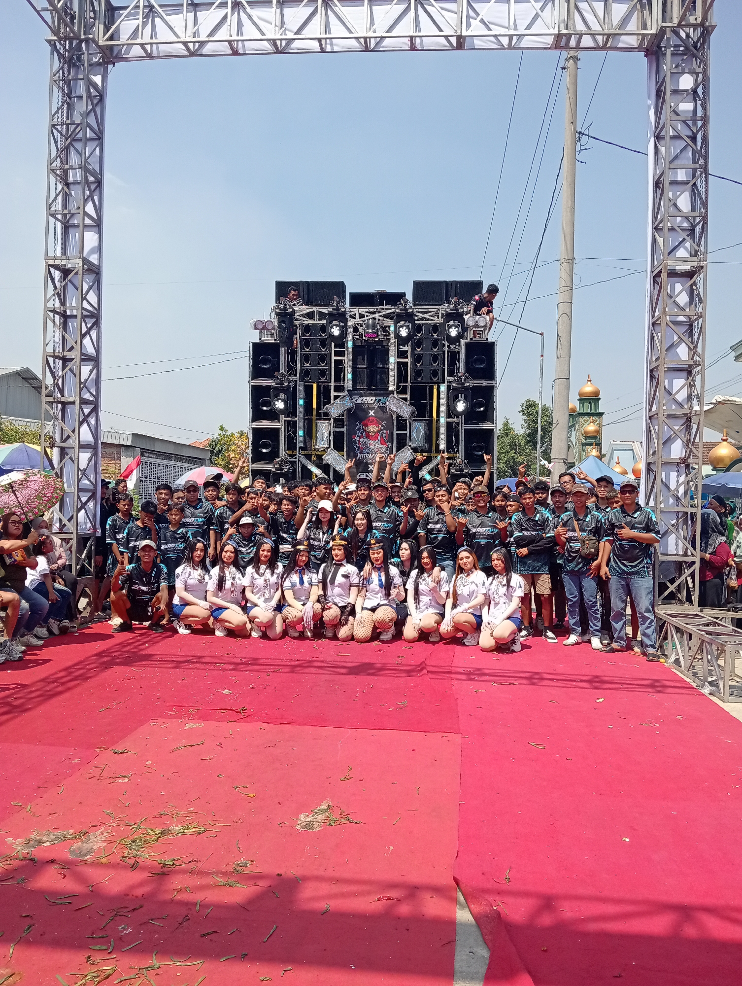 A large group of people, predominantly young adults and teenagers, are posing for a photo in front of a substantial sound system setup. The group is comprised of both males and females, with some females in the front row wearing matching outfits that appear to be uniforms or costumes. The men are mostly dressed in matching t-shirts. The sound system consists of numerous speakers stacked vertically, adorned with lights. The setting is outdoors, under a clear blue sky, with a red carpet laid out on the ground where the group is positioned. In the background, there are indications of a residential area with houses and a prominent mosque with golden domes, suggesting the location is in Wilalung, Indonesia. Metal truss structures are visible on either side, framing the scene, likely part of a stage or event setup. Several people are seated on chairs to the left of the main group, and some are standing further back. The ground on the red carpet is littered with dry grass and debris.