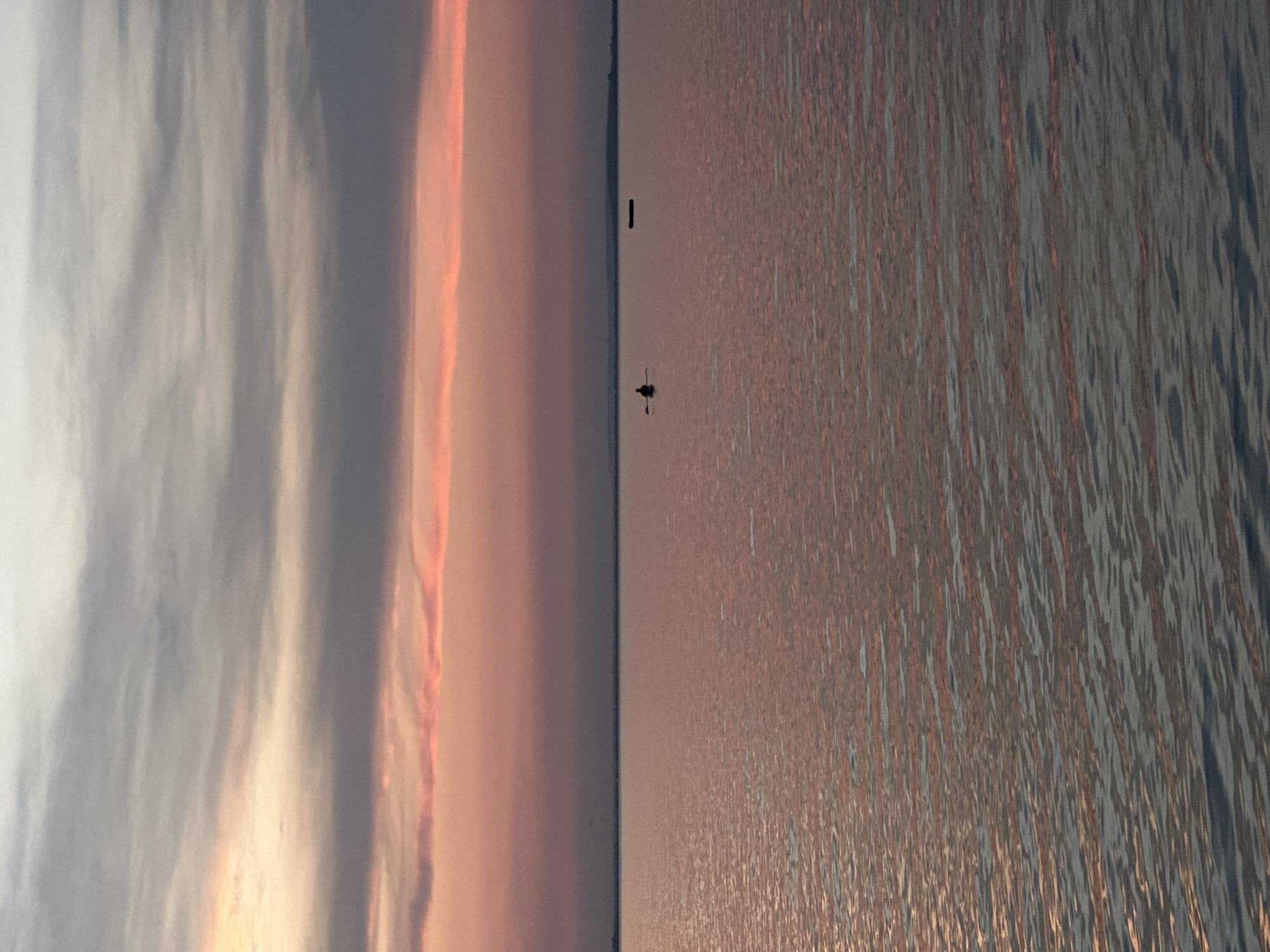 The image depicts a serene scene at dusk, likely in Kokshetau, Kazakhstan. The horizon is clearly divided between a vast sky filled with soft, wispy clouds and a calm body of water reflecting the warm hues of the setting sun. The sky transitions from a pale, cool grey on the left to shades of pink and orange on the right, suggesting the final moments of daylight. The water, in contrast, is a textured expanse of shimmering ripples, reflecting the sky's colors in muted tones of pink and grey.
In the distance, a single, small boat with two figures can be seen navigating the water. One figure appears to be paddling, adding a sense of gentle movement to the otherwise still composition. Further out, a solitary black marker or buoy stands upright in the water, a stark vertical element against the horizontal expanse. The overall mood is peaceful and tranquil, evoking a sense of quiet solitude and the beauty of nature at the end of the day. There is no visible text in the image.