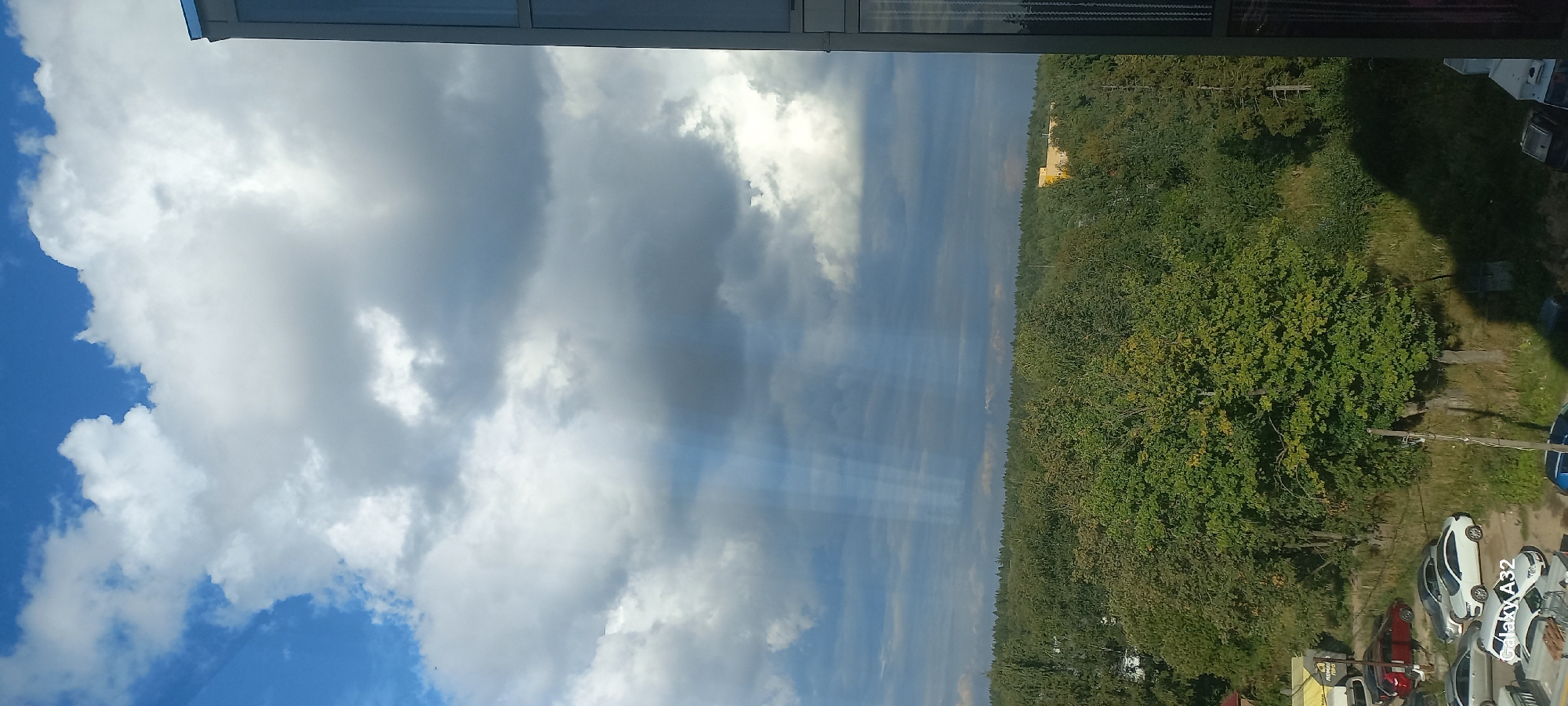 The image captures a view from an elevated position, looking out over a densely forested landscape beneath a dramatic sky. The sky is filled with a mixture of dark, brooding storm clouds and bright, fluffy white clouds, with visible sunbeams piercing through the darker masses. The foreground shows a partial view of a modern building on the right side, with a dark, reflective surface. Below the tree line, a parking area with several cars is visible, including a red car, a white car, and a silver car. The foliage of the trees is predominantly green, suggesting it is either summer or early autumn. A yellow building or structure is faintly visible in the distance, nestled within the trees on a slight elevation. The overall scene suggests a suburban or semi-rural setting on the outskirts of Elino, Russia, with a mix of natural and built environments.