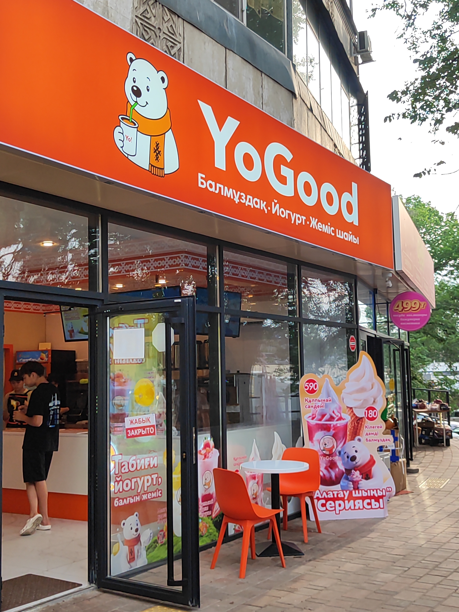 The image shows the exterior of a "YoGood" frozen yogurt shop located on a street in Almaty, Kazakhstan. The storefront has a prominent orange awning with the "YoGood" logo, which features a cartoon polar bear drinking from a cup. Below the awning, large glass windows display the interior of the shop, which appears to be brightly lit. Inside, customers are visible, as well as displays of yogurt and frozen desserts.
On the sidewalk in front of the shop, there are outdoor seating arrangements with bright orange chairs and white tables. A large promotional sign stands near the entrance, showcasing different ice cream and yogurt products. This sign, and other signage on the shop's doors and windows, are written in a combination of Kazakh and Russian, as evidenced by words like "ЖАБЫҚ ЗАКРЫТО" (Closed) and prices like "590" and "180" alongside descriptions of the items. A price of "499т" is also visible on a circular pink sign hanging from the awning. The overall scene suggests a commercial area with pedestrian traffic. The architecture of the building above the awning appears to be typical of urban Almaty, with concrete and decorative elements. Trees are visible in the background, suggesting an outdoor urban environment.