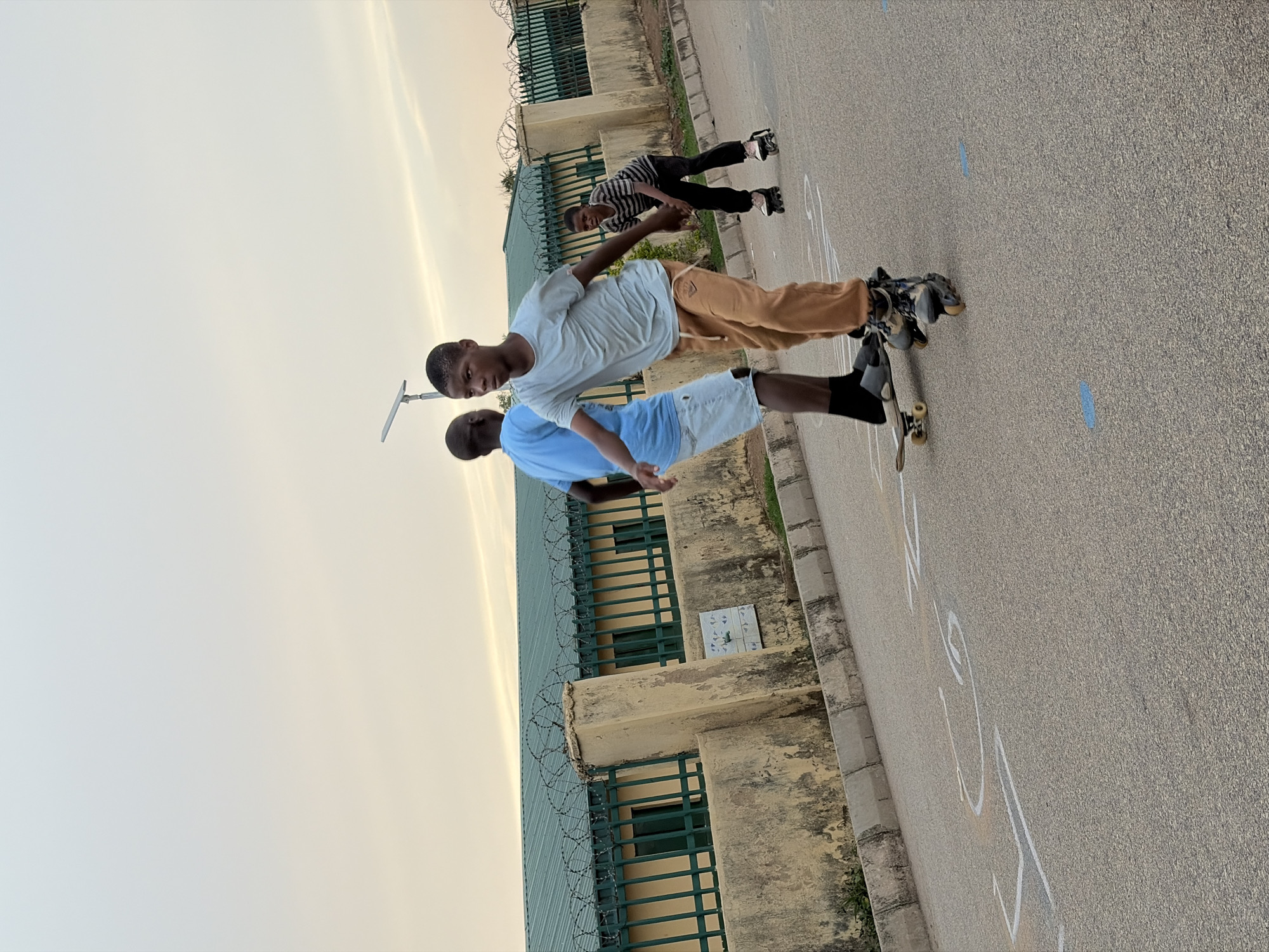 The image depicts three boys in what appears to be an outdoor area near a building, potentially in Jos, Nigeria, though the exact location cannot be confirmed without further details.
The first boy, in the foreground, wears a white shirt and brown pants. He is standing on a skateboard wearing rollerblades. A second boy, wearing a blue shirt and white shorts, is standing behind him. A third boy, wearing a striped shirt and black pants, is further in the background.
The setting includes a building with a green roof, green bars over the windows, and barbed wire on top of the structure. The ground is paved. White lines are drawn on the ground. The sky appears light, possibly indicating dawn or dusk.
