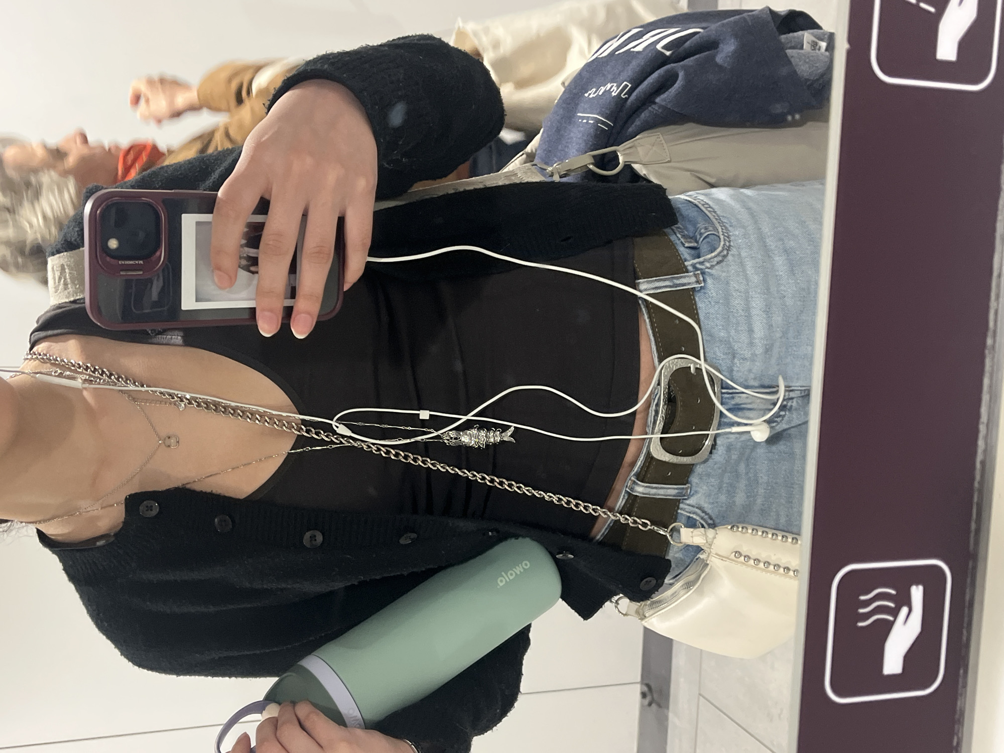 The image shows a person's selfie in a mirror. The person is wearing a black cardigan over a black top, blue jeans with a brown belt, and several silver necklaces. They are holding a cell phone in one hand, a green water bottle in the other, and wearing white earphones. There is a white purse with silver studs hanging from their belt. The reflection in the mirror shows other people in the background. In the foreground, there is a sign with a white hand over curved lines, on a purple background. It's unclear if this photo was taken in Colombier-Saugnieu, France, as no clear landmarks or signs indicating the location are visible in the image.