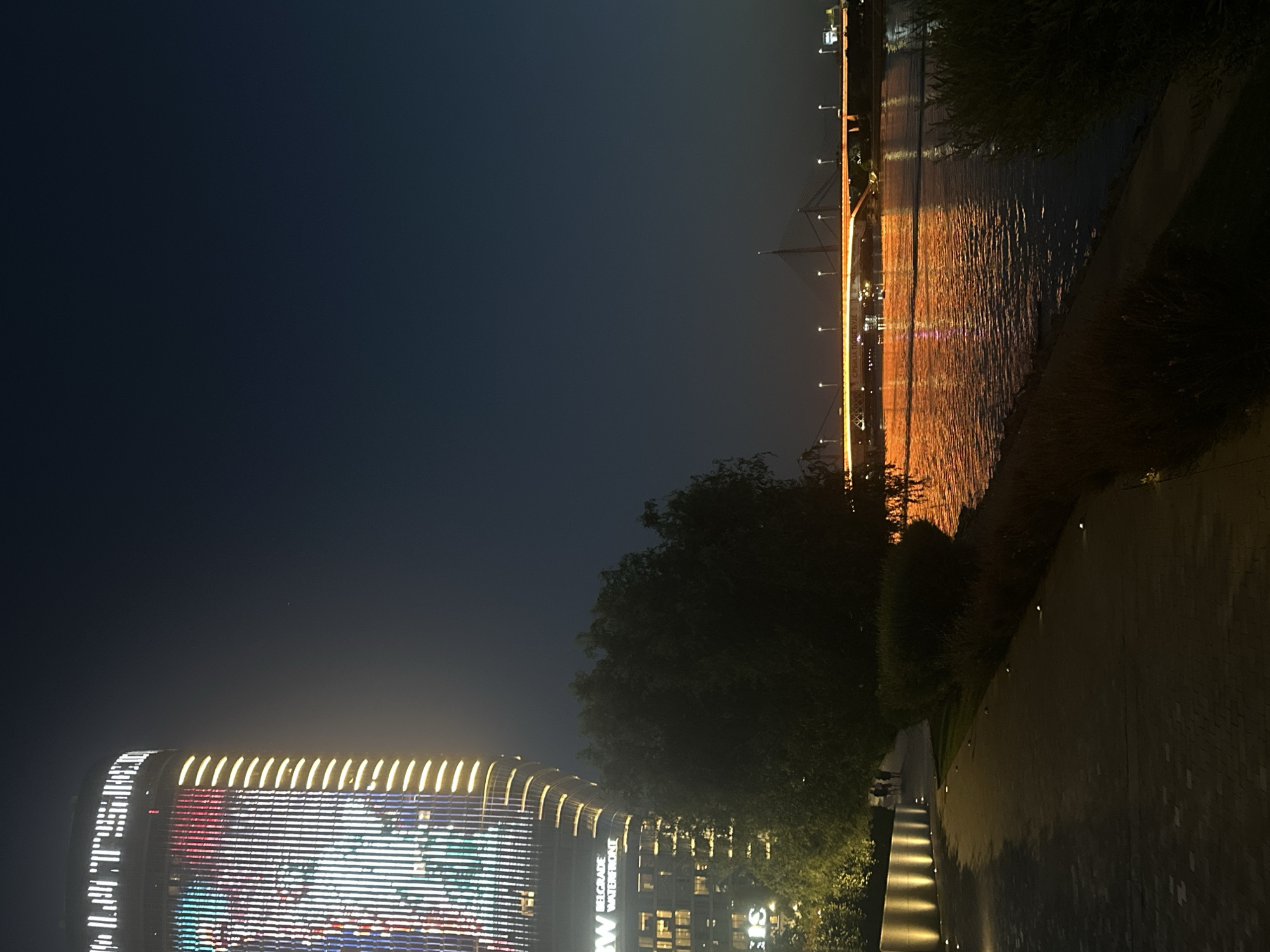 The image captures a nighttime scene along the Sava River in Belgrade, Serbia, likely near the Belgrade Waterfront. On the left, a modern, illuminated skyscraper, identified by the text "BELGRADE WATERFRONT" on its facade, stands tall. The building features a dynamic facade with vertical lines of light in various colors, creating a vibrant display against the dark sky. Adjacent to the skyscraper is a pathway illuminated by ground-level lights, with sparse vegetation and trees flanking the riverbank. In the distance, a bridge, possibly the Ada Bridge, is brightly lit with orange lights, its structure visible against the deep blue night sky. The river reflects the city lights, creating a shimmering orange and red glow on its surface. The overall atmosphere is urban and contemporary, illuminated by artificial lights against the natural darkness.