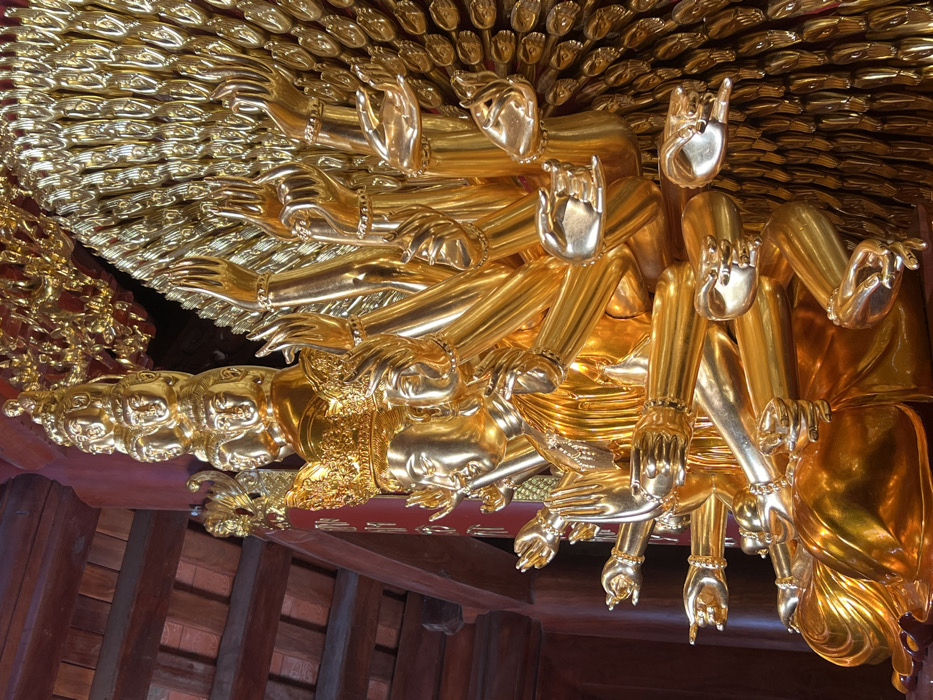 A close-up shot showcases a golden statue, possibly of a deity, adorned with numerous arms and hands extending outwards in a fan-like formation. The intricate details of the hands, including bracelets, are visible. The statue is positioned near a wooden structure, possibly a building or temple interior, evident from the dark wood beams in the lower portion of the frame. The golden color dominates the image, reflecting light and highlighting the statue's ornate design.