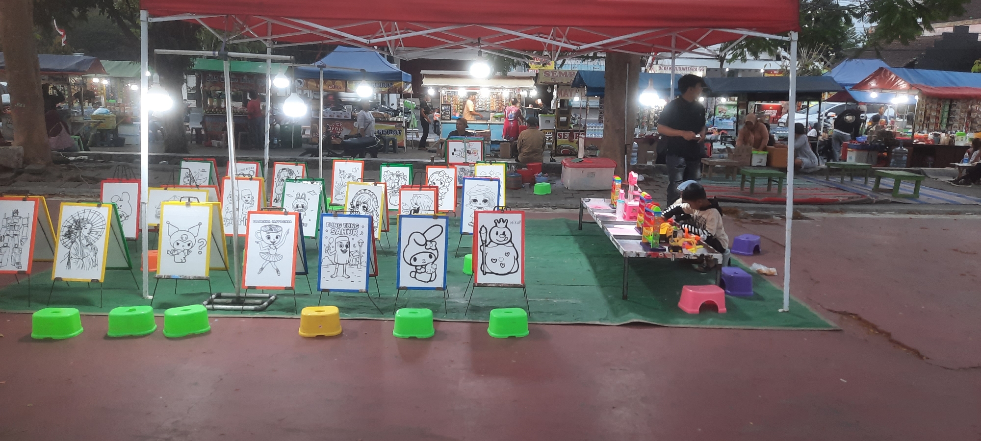 This image depicts an outdoor scene in Kanigoro, Indonesia, likely a market or fairground. In the foreground, an assortment of white drawing boards with black and white line art are displayed on easels. These drawings feature various characters, including robots, anime-style figures, and animals, suggesting they are intended for coloring activities. Three bright green plastic stools are placed in front of some of these boards. A child is seated at a long table on the right, engaged in playing with colorful toys. Several other children's stools, in purple and pink, are scattered around the table. Behind the drawing boards, a large red canopy tent covers part of the area. Further in the background, various market stalls are visible, some with awnings and lighting, indicating commercial activity. People can be seen browsing or working at these stalls. The ground is paved with a reddish-brown surface, with green mats laid out in some areas, particularly around the drawing boards and the table with toys. The overall atmosphere suggests a lively, family-friendly event.