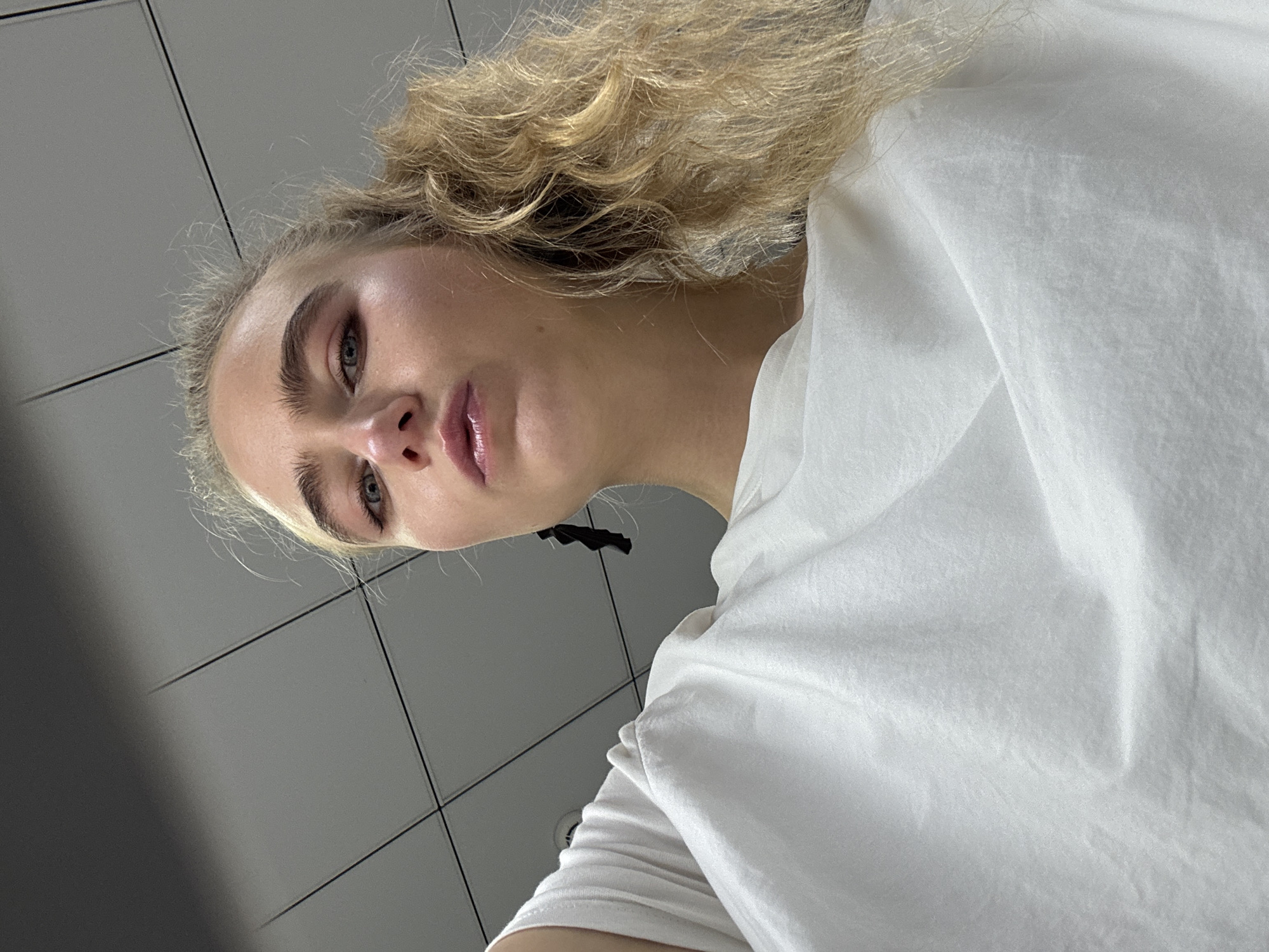 A low-angle shot depicts a fair-skinned person with blonde, wavy hair and defined eyebrows. The person's eyes are directed towards the camera. They are wearing a white T-shirt. The background features gray tiled walls. The location is unknown.