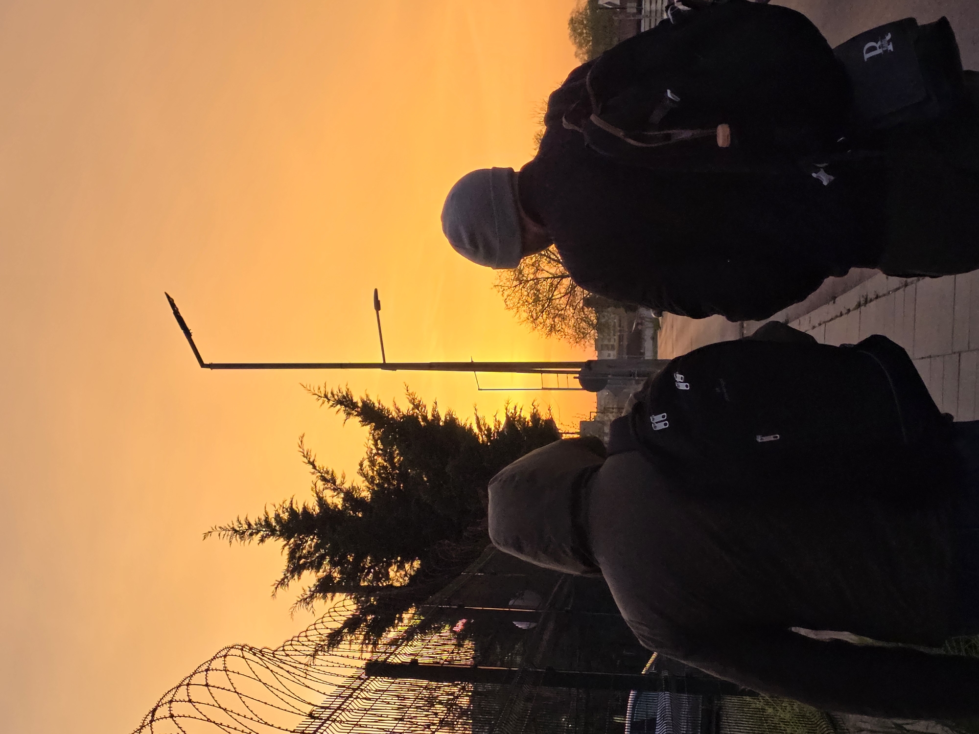 Two people, possibly male, are silhouetted against an orange sky, likely during sunset or sunrise. The individual in the foreground wears a hooded jacket and a backpack. The person behind wears a dark jacket and a beanie. Both appear to be observing something in front of them. In the background, there's a barbed wire fence and a tree with sparse foliage. There are also lampposts visible. Based on the environment and the direction of the sunset/sunrise, the location is possibly near the Maritsa river, but that cannot be stated with certainty.