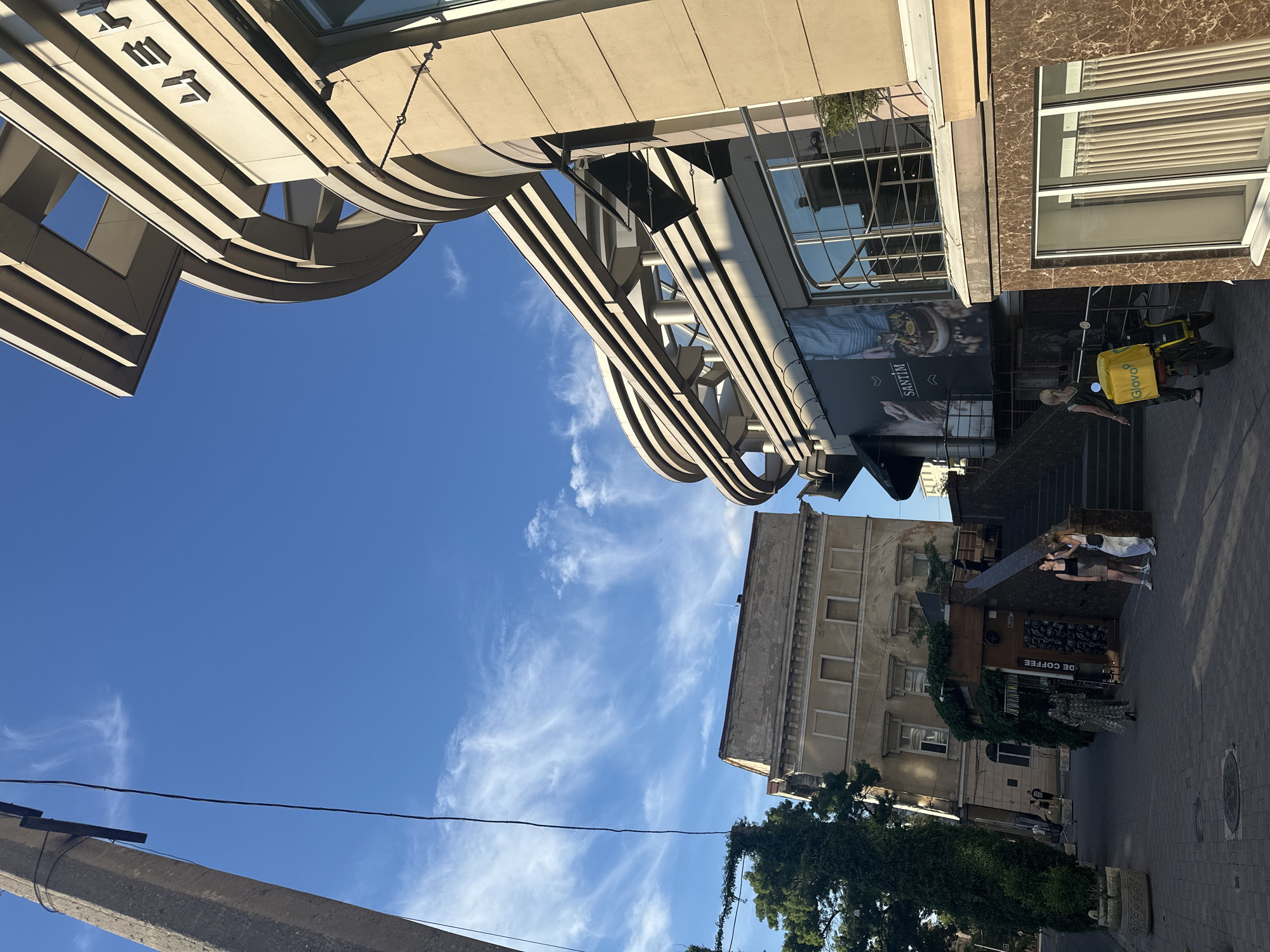 The image shows a street scene in Odesa, Ukraine, on a clear day with scattered white clouds in a bright blue sky. The perspective is looking upwards, with architectural elements of buildings dominating the upper portion of the frame.
On the left, a curved, beige building with layered horizontal accents is visible, with large letters spelling out "TER" at the top. Adjacent to it, a building with a marble facade and blinds on its windows is also present.
In the middle ground, a street with cobblestone paving is visible. A person wearing a yellow "Glovo" delivery bag on a scooter is parked on the right side of the street, facing away from the viewer. They are wearing a dark t-shirt and pants. Two other people are standing on steps to the right, appearing to converse. One person is wearing a black tank top and plaid skirt, while the other has a white top and blue shorts.
A building with a weathered facade and multiple windows is situated behind the scooter. Below this building, a storefront with the words "DE COFFEE" is visible, adorned with ivy and green foliage.
Further down the street, more people are visible, including a person in a patterned dress. A large tree with dense foliage is on the left side of the street, and another tree with a similar style of pot is at the bottom left.
The sunlight suggests it is daytime. The overall atmosphere appears to be a typical urban environment.