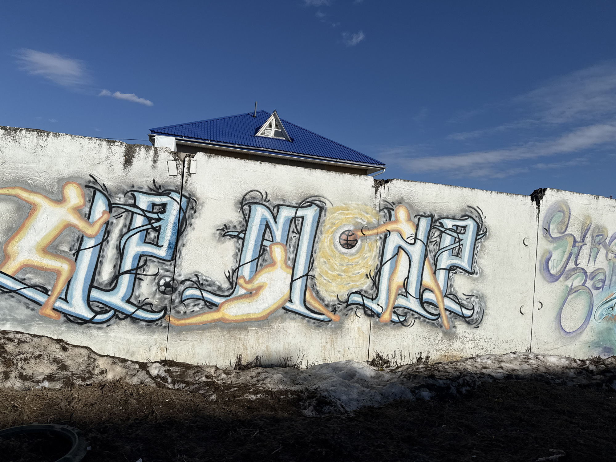 The image shows a section of a graffiti-covered white wall with a blue-roofed structure visible behind it, set against a clear blue sky. The wall is adorned with stylized graffiti art incorporating figures and text. Patches of snow and dark soil are visible at the base of the wall. The location is in the city of Unknown, Unknown.