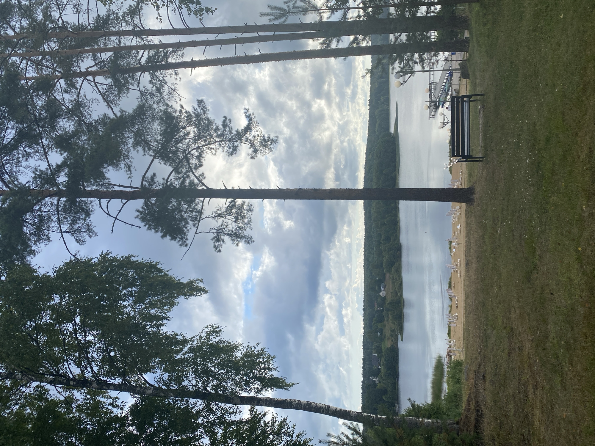 The image depicts a tranquil lakeside scene at Krasnoflotskoe, Russia, likely in the early evening or on an overcast day. In the foreground, a dark wooden bench sits on a grassy knoll, offering a vantage point overlooking the lake. Beyond the bench, a sandy beach is lined with numerous white lounge chairs and umbrellas, suggesting a recreational area. A small wooden pier extends into the calm, gray water, with several boats moored alongside.
The middle ground is dominated by the expansive lake, its surface reflecting the cloudy sky. Across the lake, a dense, verdant forest forms a natural border, with scattered houses and cottages nestled among the trees, indicating a small settlement. The sky is filled with dramatic, grey clouds, with patches of lighter sky suggesting the sun is attempting to break through.
The foreground also features several tall trees, including a birch tree on the left with its distinctive white bark and a cluster of pine trees on the right. The overall impression is one of peaceful seclusion and natural beauty.