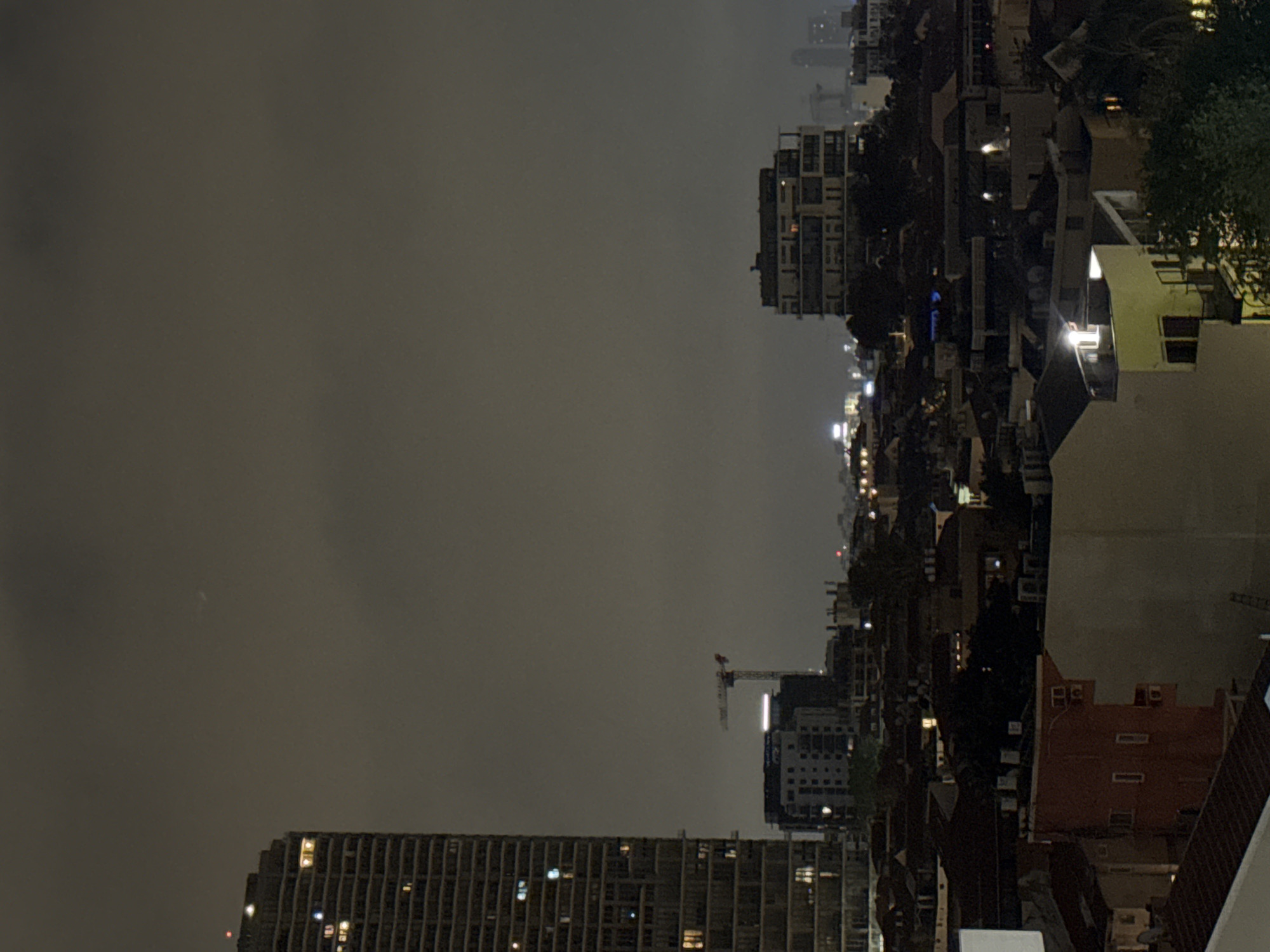 The image captures a night scene in Tel Aviv, Israel, under a heavily clouded sky. Predominantly featuring a cityscape with a mix of residential buildings and high-rise structures, the photo is oriented with the sky occupying the left side. Buildings with lit windows are visible, indicating nighttime. A construction crane is present among the buildings. The overall light level is low, with illumination mainly from artificial sources within the urban environment.