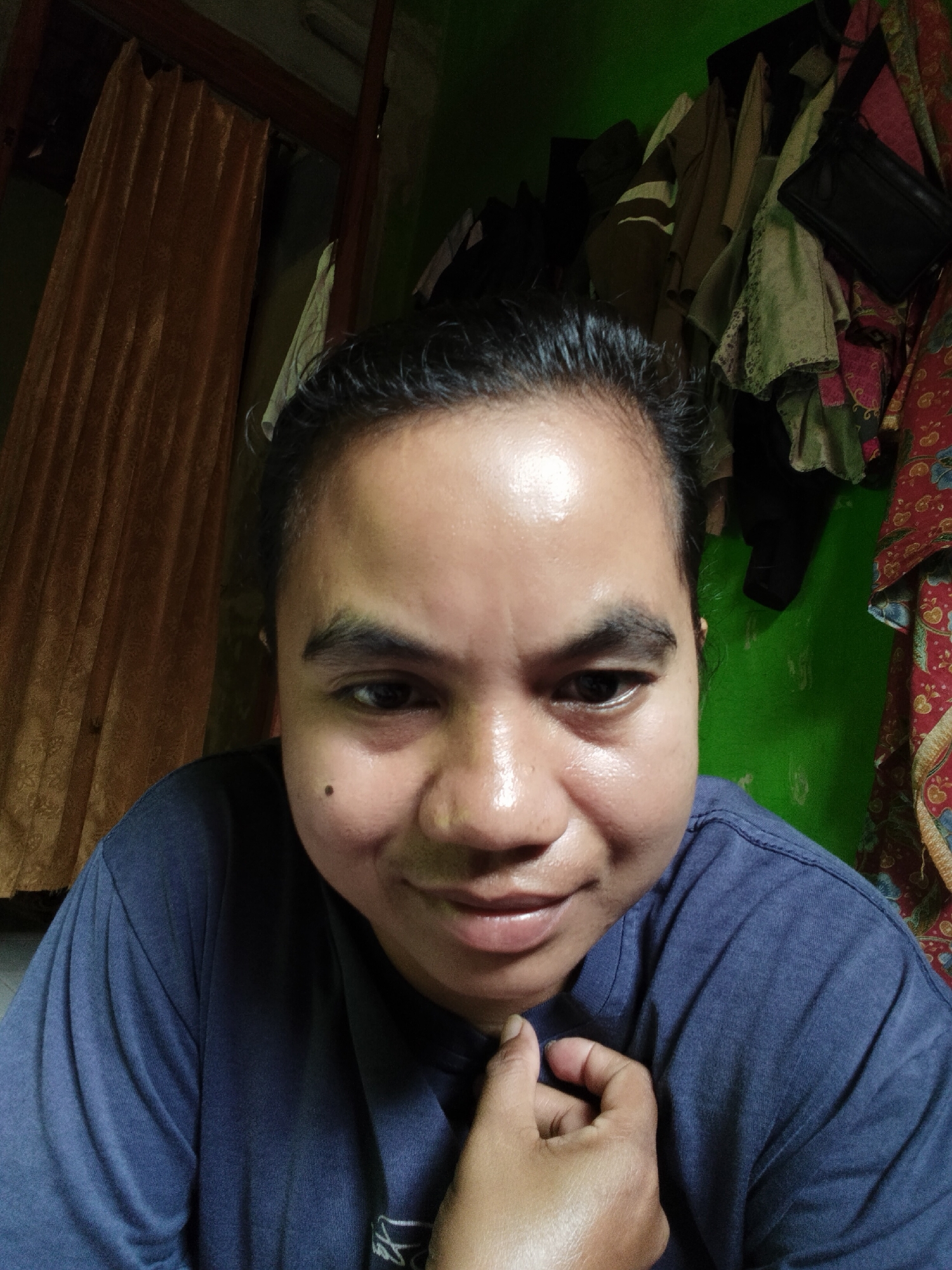In Tanjung Harta, Indonesia, an individual with dark, slicked-back hair is captured in a close-up, selfie-style portrait. They are wearing a dark blue t-shirt, and their right hand is positioned near their chest, with fingers lightly touching the fabric. The individual has a mole on their left cheek. The background features a green wall, with a collection of clothing hanging to the right. To the left, a section of a textured, golden curtain is visible. The lighting highlights the shine on the individual's forehead and cheeks.