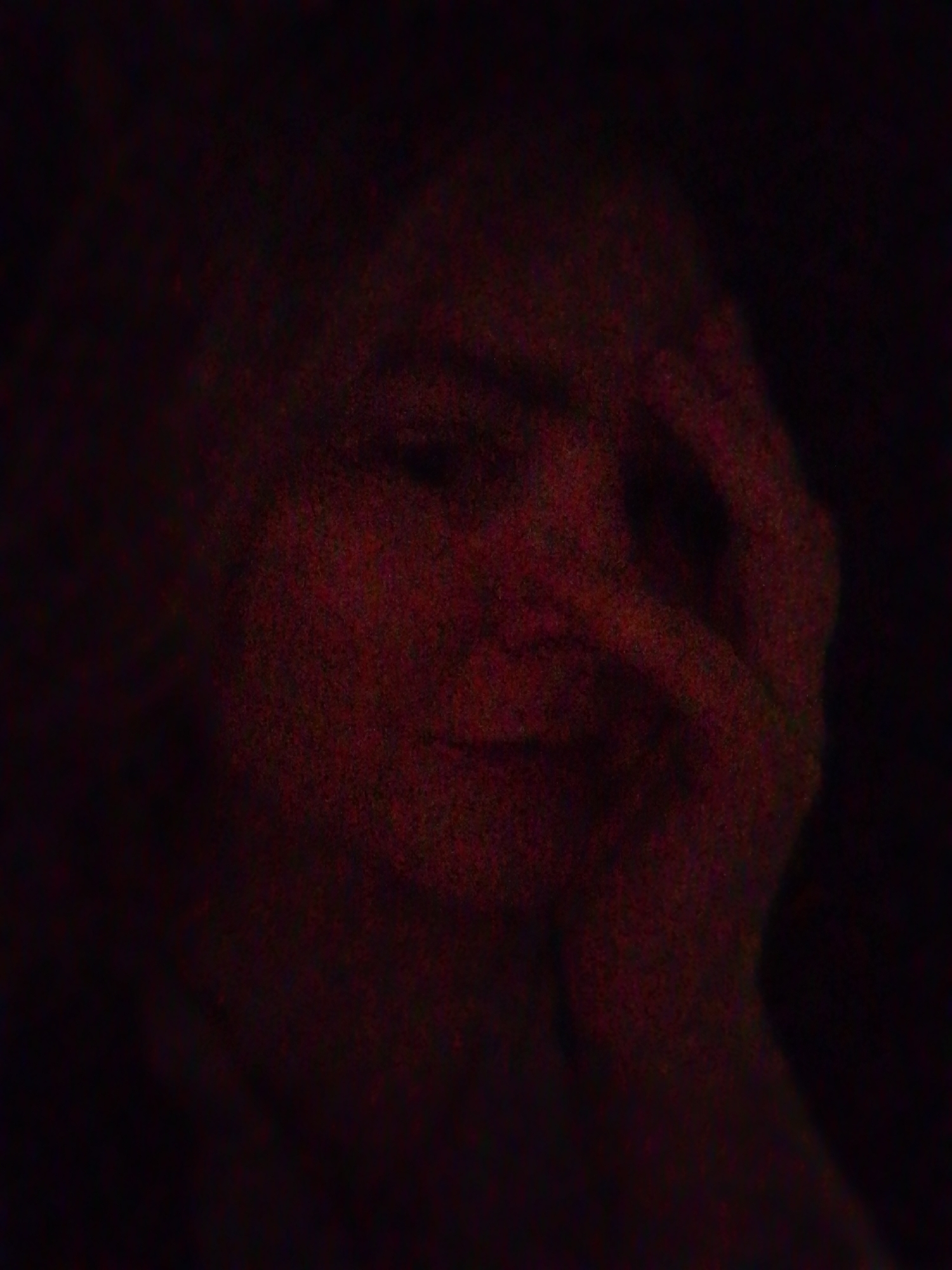 The image is a low-light, grainy depiction of a person's face with a hand partially obscuring it. The scene is dark, and details are indistinct due to the poor lighting conditions. The individual's features are barely discernible, but it appears to be a face in repose, with the fingers of one hand touching the face. The overall color tone is predominantly reddish-brown due to the limited light source. There is no indication of the location being Izhevsk, Russia, or any other specific geographic place. There are no identifiable objects or actions beyond the aforementioned description.