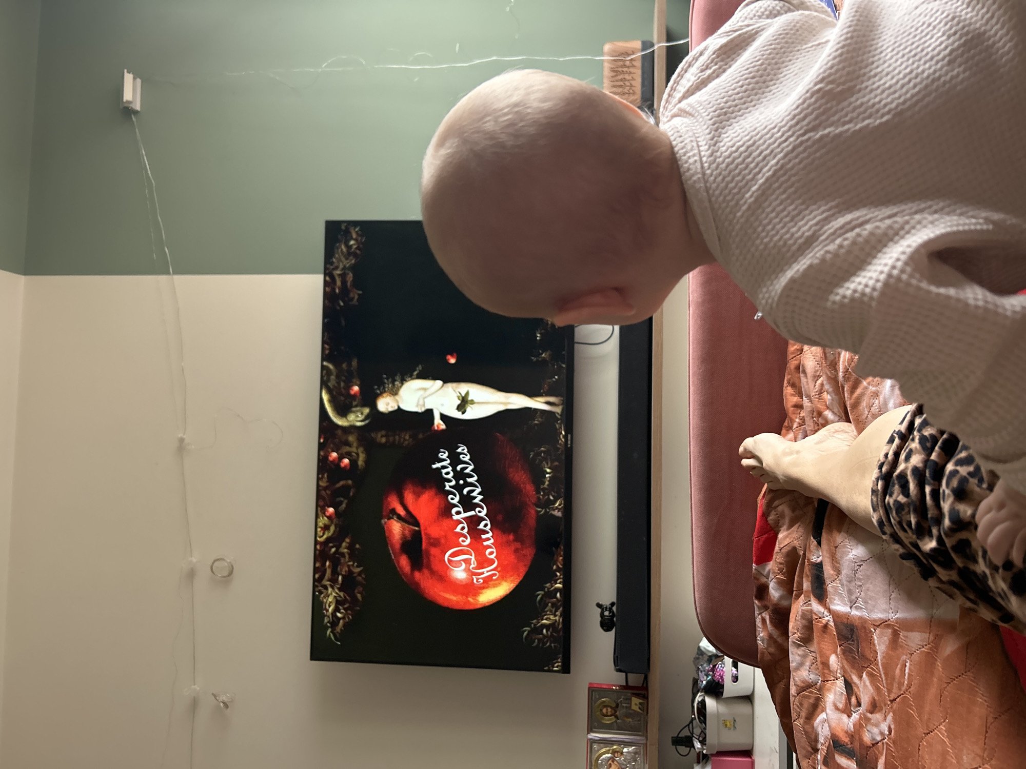 A close-up, slightly tilted indoor shot shows a baby with a bald head and wearing a white, textured garment, leaning over a scene. The scene includes a television screen displaying cover art for the TV series "Desperate Housewives," featuring an apple and a woman. Behind the baby is a pink object, likely part of furniture. A leg and foot with an animal print fabric visible rest on a brown and red patterned surface. The wall is two-toned, with white below and green above, and electrical wires run down the wall. The location cannot be determined from this image. The TV show title is English, however, indicating that this scene is unlikely to be in Russia.