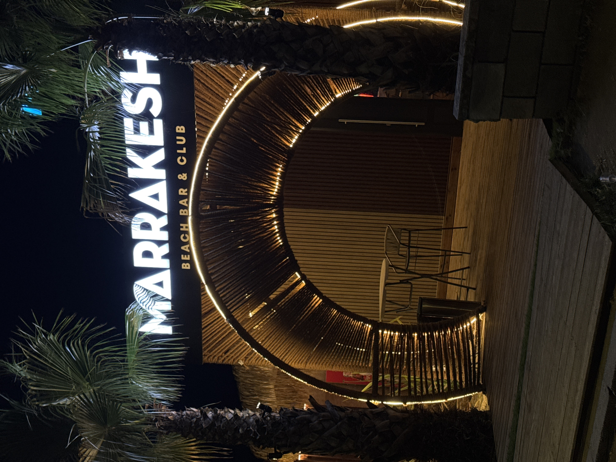 The image shows the entrance to "MARRAKESH BEACH BAR & CLUB" in Burgas, Bulgaria at night. The entrance is framed by a large, arched structure made of interwoven bamboo or reeds, illuminated by warm-toned LED strip lights. Two tall palm trees flank the entrance, their fronds silhouetted against the dark sky. The sign for "MARRAKESH" is illuminated with white LED lights and is positioned above the arched entrance, with "BEACH BAR & CLUB" written in smaller letters below. Inside the archway, a small round table with two metal chairs is visible on a wooden deck. A dark-colored cylindrical trash can is next to the table. The building behind the entrance appears to have a wooden facade. The foreground consists of a wooden walkway. The overall atmosphere is tropical and inviting, characteristic of a beach club.