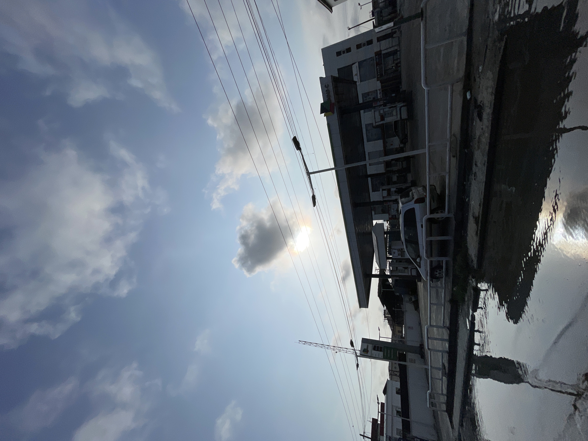 This is a wide-angle, low-angle shot of a gas station in Lekki, Nigeria, under a partly cloudy sky. The sun is visible through a large cloud formation, casting a bright halo. Overhead power lines crisscross the sky.
In the foreground, a large puddle of murky water reflects the sky and surrounding structures. A white metal railing runs along the edge of the puddle. Beyond the railing, a silver SUV is parked under the gas station's canopy. The gas station itself is a modern, multi-story building with a prominent white facade and a large, flat canopy supported by pillars. Several gas pumps are visible under the canopy. A tall pole with a cellular antenna stands to the left of the gas station.
The scene appears to be at dusk or dawn, given the lighting conditions and the presence of streetlights on poles. There are a few figures of people visible within the gas station and on the sidewalk, but they are small and indistinct. The environment suggests a typical urban setting with a mix of commercial and residential buildings.