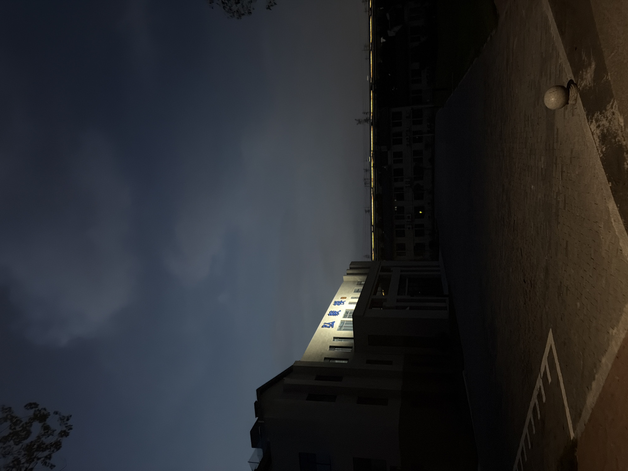 The image shows an exterior view of a modern building with blue Chinese characters illuminated on its facade. The building is constructed with light-colored material and has several windows. In front of the building, there is a paved courtyard area. The sky is dark, suggesting it is either dusk or nighttime, with some clouds visible. In the distance, another building or series of buildings can be seen with lights on. The foreground of the image shows a portion of the paved courtyard, with white markings indicating a path or parking area. A spherical stone object rests on a small stone base in the courtyard. The overall scene appears to be in a quiet urban or suburban setting. The building signage is in Chinese, and the context of the building's illuminated name on the facade suggests it could be a business, institution, or public facility. Given the provided context, the location is identified as Jiangjin, China.