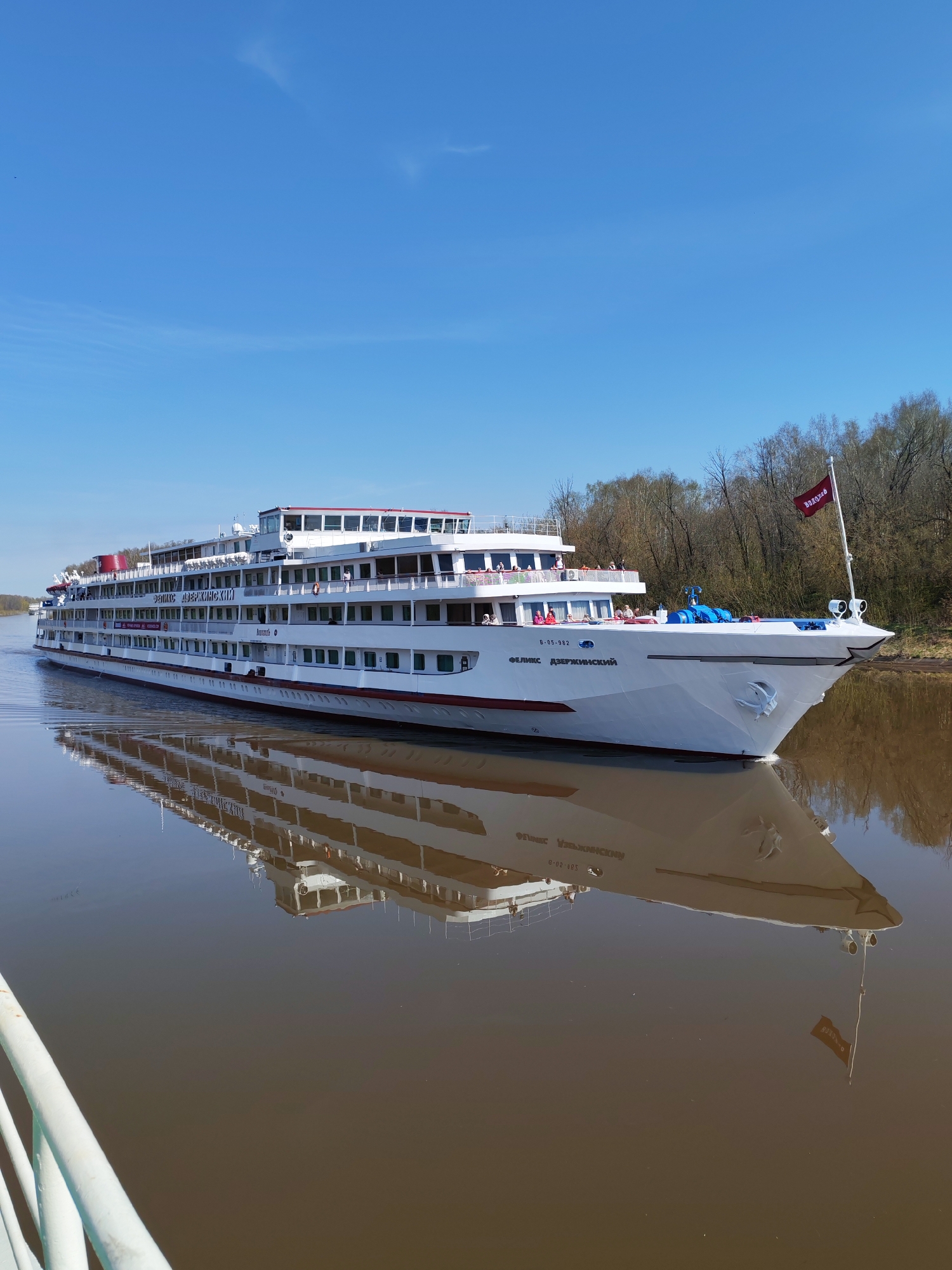 The image depicts the "Felix Dzerzhinsky" river cruise ship sailing on a river, likely the Yakhroma River in Russia. The ship is white with red and grey accents, and several decks are visible. Passengers can be seen standing on the decks. A flag with the word "Вологда" (Vologda) is flying at the bow. The ship's name is visible on the hull. The river water is brown. The sky is blue. Trees are visible along the riverbank. The ship's reflection is clear in the water. A portion of a white railing is visible in the lower left corner, suggesting the photo was taken from another boat or dock.