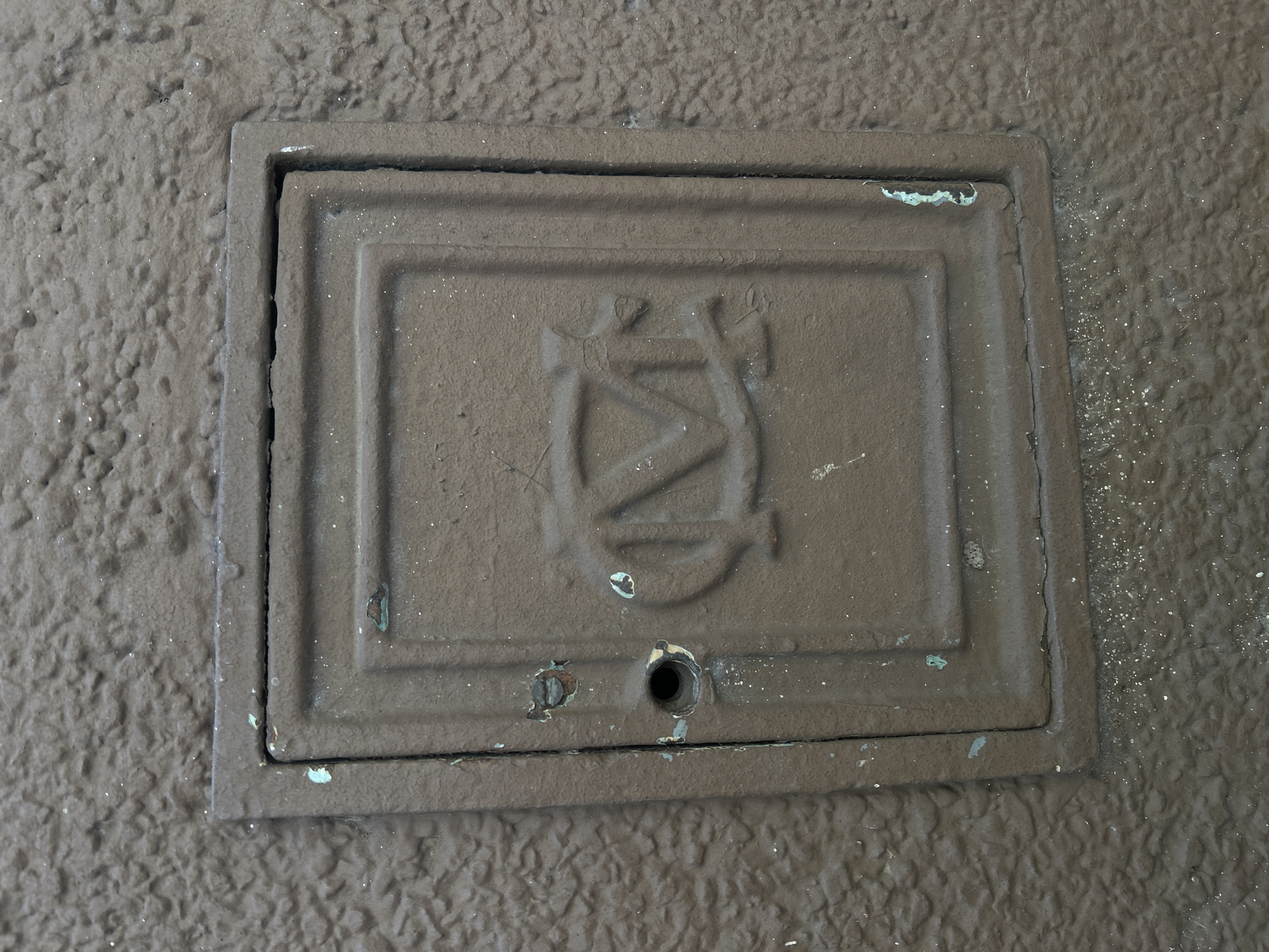 An overhead view captures a rectangular, dark brown metal utility cover or access panel set into a rough, textured brown surface, likely pavement or concrete, in Buenos Aires, Argentina. The metal cover features raised rectangular borders, creating a framed appearance.
At the center of the cover, a prominent, stylized monogram is cast in relief. This intricate design appears to incorporate intertwined letters, possibly "D" and "N" or "N" and "D," topped with a small, crown-like or decorative element. Near the bottom edge of the cover, beneath the monogram, there is a circular opening, and to its left, a corroded or damaged screw head is visible.
The cover shows significant signs of age and exposure, including areas of greenish-blue oxidation or patina, particularly along its edges and within the central design. The dark brown surface of the cover itself appears somewhat worn, with minor chips or flakes in its coating. Small, white, sparkling specks, resembling glitter or tiny debris, are scattered across both the metal cover and the surrounding bumpy surface. No specific activity is taking place; the image is a static shot of this urban fixture.