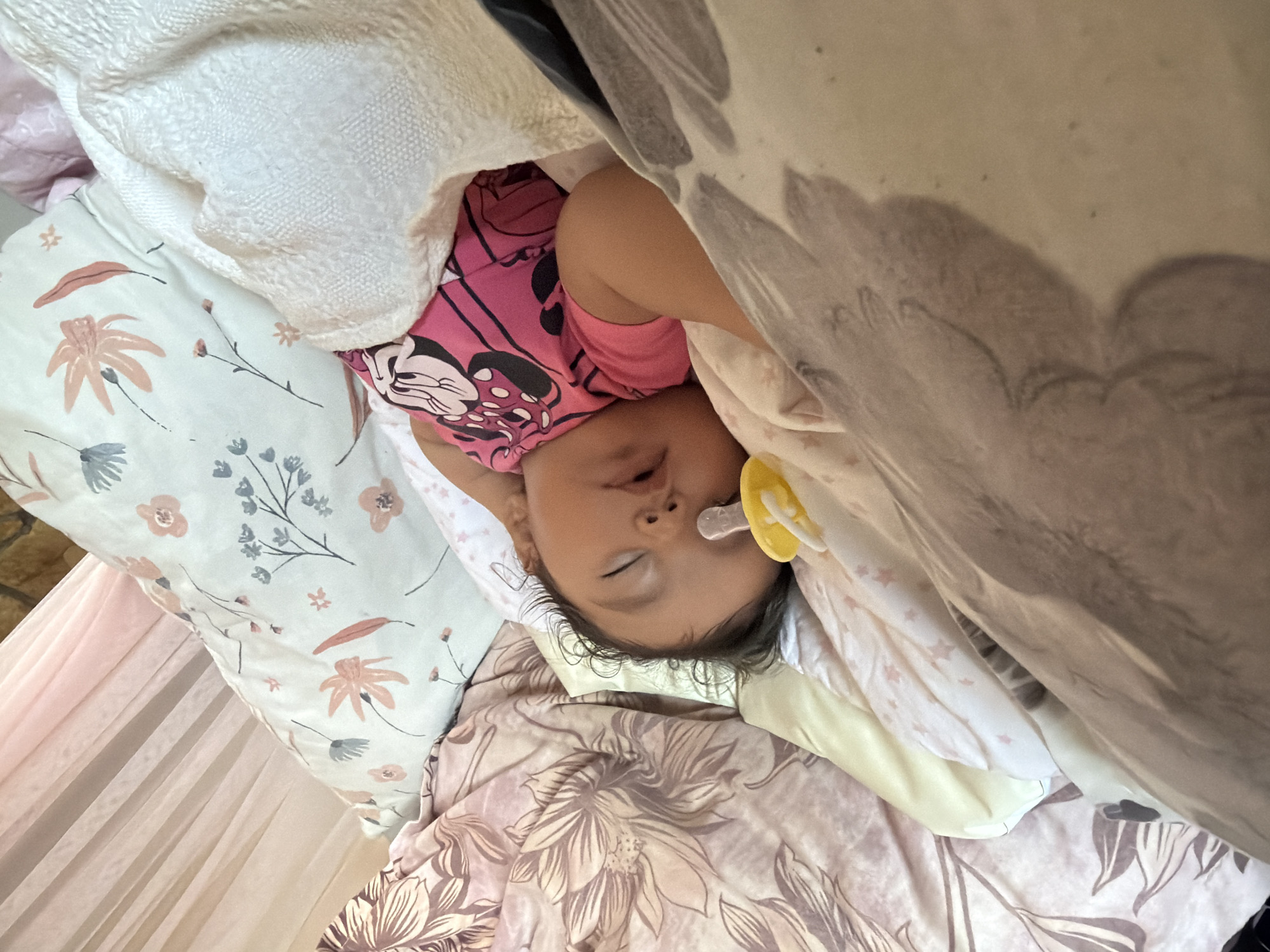 The image shows a baby sleeping on a bed. The baby is wearing a pink shirt with Mickey Mouse printed on it and has a pacifier in its mouth. The bed is covered with floral patterned sheets and a white blanket. There are several pillows on the bed, one with a floral pattern and another with a light blue background and floral designs. A sheer pink curtain is visible in the background. The scene appears to be indoors, likely in a bedroom. The provided location of Razhevo Konare, Bulgaria is not discernible from the image itself.