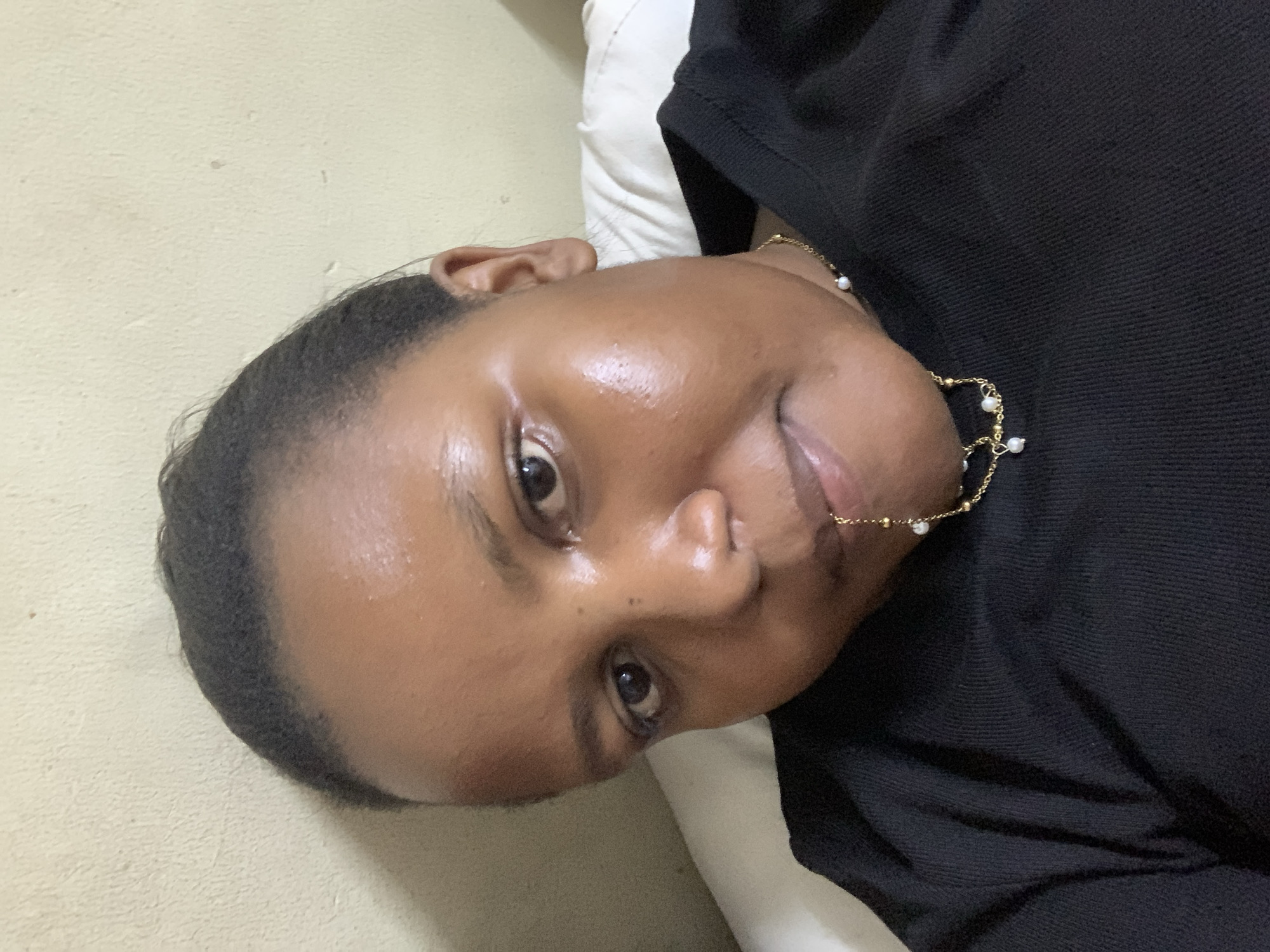 A close-up photograph shows a person with dark skin, likely female, with short, dark hair. They are wearing a black garment and a delicate gold necklace with small white pearls. The person is lying down, with their head resting on a white pillow or cushion. The background is a plain, off-white wall. Without additional information, specifying the location within Dar es-Salaam is impossible.