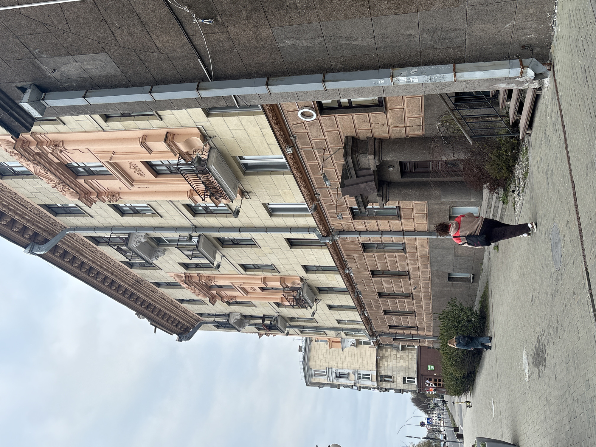 The image, taken in Minsk, Belarus, presents an urban street scene featuring a row of multi-story buildings and two individuals on a wide sidewalk under an overcast sky.
The prominent building on the left is characterized by its cream and light-pink facade, adorned with classical architectural elements such as pilasters, cornices, and decorative moldings. Numerous windows are visible, with several air conditioning units installed beneath them. A grey metal downspout runs vertically along this building's exterior.
Adjacent to it, a second building displays a darker, possibly brown or grey, facade with a distinct textured, block-like pattern. This structure also incorporates decorative features, including what appear to be large columns at its lower levels. Another metal downspout is visible along its upper edge, bearing some indistinct writing or graffiti. In the distance, further buildings line the street, contributing to the urban panorama.
On the paved sidewalk, two people are discernible. Closer to the viewer, one individual is seen from behind, wearing dark pants, white sneakers, and a two-toned jacket, beige on top and red/orange on the bottom. They are carrying a dark bag over their shoulder. Further down the sidewalk, near a cluster of green bushes, another person in blue jeans and a light-colored top is partially visible, seemingly bending down.
The sidewalk itself is composed of patterned concrete pavers and features manhole covers. Beyond the immediate foreground, a street is visible, with parked cars, streetlights, and traffic lights. A small sign with the number "50" is attached to a distant building. The sky is uniformly grey and overcast, indicating a lack of direct sunlight, suggesting a cool or mild daytime environment.