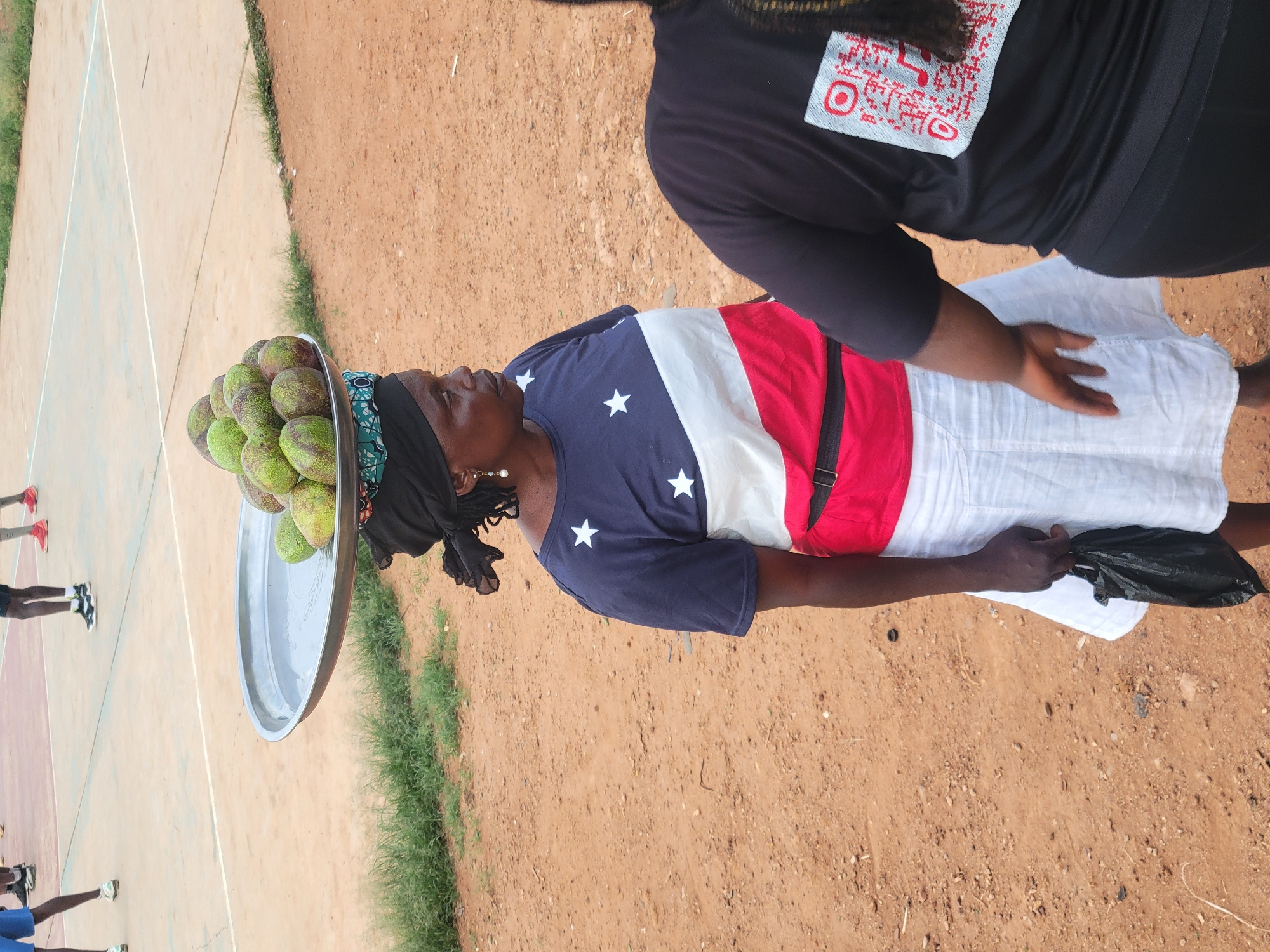 A woman balances a silver tray laden with numerous green fruits atop her head, standing on a reddish-brown, dusty surface in Jos, Nigeria. She wears a head covering, a blue t-shirt with white stars, a white panel, and a red panel with a black strap. She holds a black plastic bag in her left hand. Another person, partially visible to the right, wears a black shirt with a white design on the back. A paved surface with court lines and figures of people are visible in the distance.