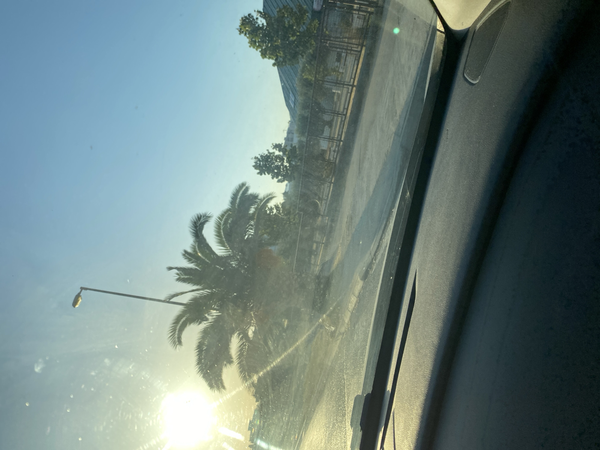 The image depicts a view from inside a vehicle looking out through the windshield. The sun is bright and low in the sky, casting strong rays that create lens flare and illuminate the scene with a warm, golden light. A palm tree is prominently featured, its fronds silhouetted against the sky. In the background, a street light is visible. Beyond the palm tree, there is a street with a sidewalk and a fence. Several trees, including olive trees, are planted behind the fence, and a building with a green roof is situated further back. The foreground shows the dashboard of the car. The sky is clear and blue.
The scene appears to be captured in an outdoor, urban or suburban environment with vegetation and infrastructure. Based on the visual cues of palm trees, clear blue sky, and the general aesthetic, the location could be a coastal city in a Mediterranean climate. Given the request to include the location in the city of Bar, Montenegro, the scene is likely situated in Bar, Montenegro, a coastal city known for its palm-lined streets and pleasant climate, especially during times when the sun is low in the sky as depicted.