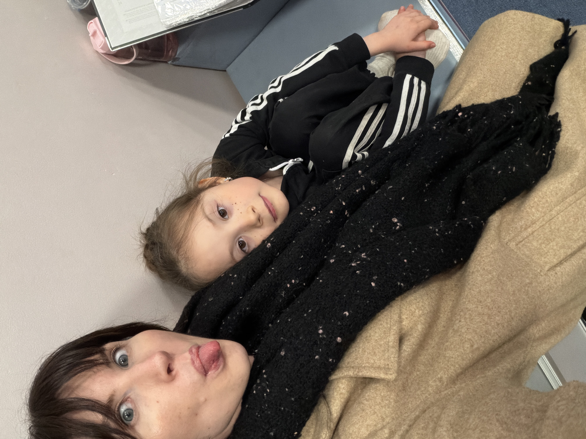 This indoor selfie captures an adult woman and a young child relaxing together in what appears to be a casual public or waiting area in Perm, Russia. The image is rotated, suggesting a shot taken while lying down or leaning back.
In the foreground, an adult woman with dark hair and blue eyes makes a playful expression, sticking out her tongue and widening her eyes. She is wearing a light tan or beige coat and a thick, black textured scarf with small, lighter flecks.
Behind her, a young child with light brown hair and a scattering of freckles on their nose looks towards the camera with a calm, curious gaze. The child is dressed in a black tracksuit with distinctive white stripes along the arms and legs, reminiscent of Adidas athletic wear, and white socks.
The background is minimal, featuring a plain light grey surface and a darker blue upholstered surface, possibly a bench or sofa. Some papers or files are faintly visible in the upper background, but no text is clearly legible. The overall impression is one of a lighthearted and affectionate moment shared between the two individuals.