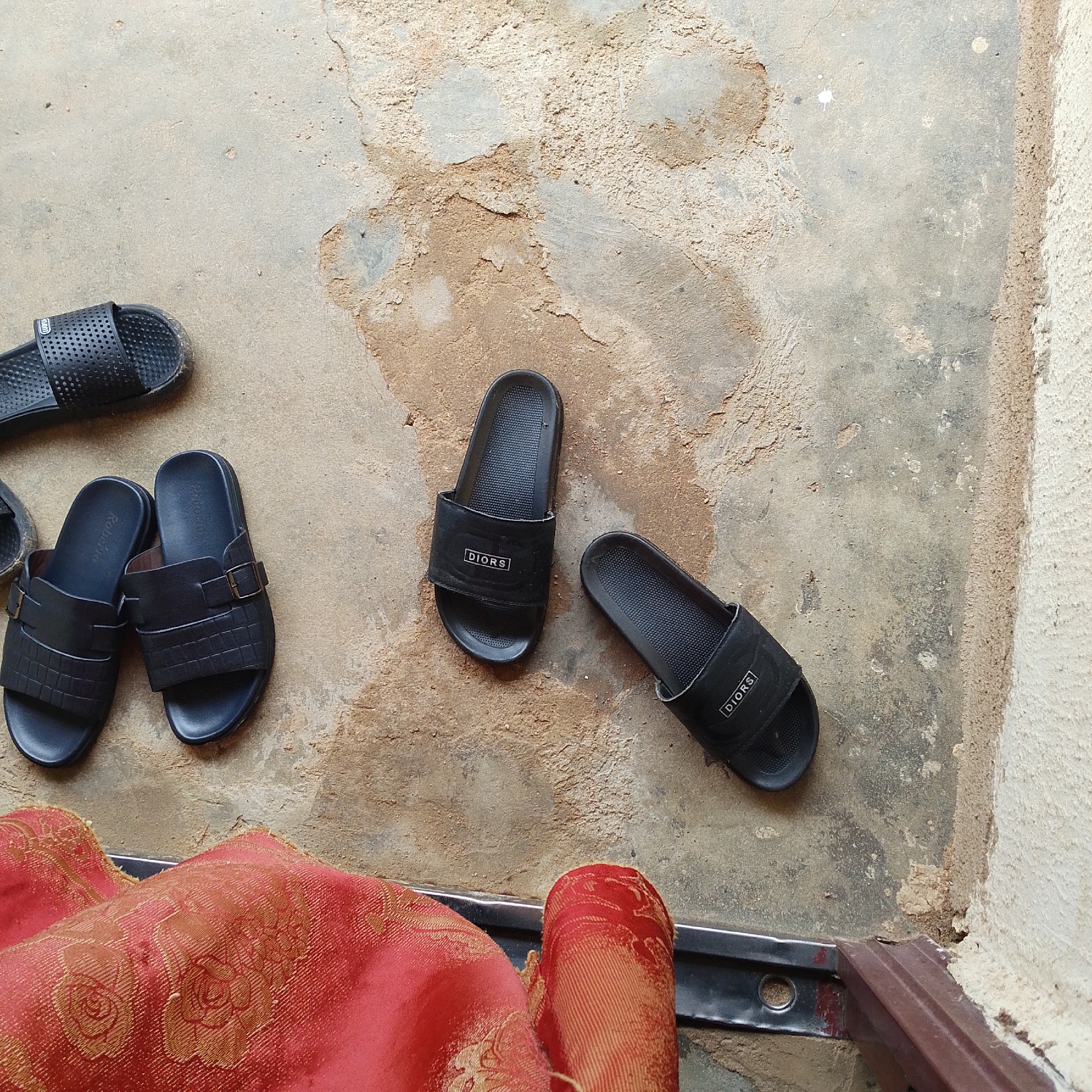 The image displays a top-down view of several pairs of black sandals on a textured, light-colored concrete surface. The sandals are scattered and appear to be on an outdoor ground, possibly near a doorway or walkway. In the foreground, a red fabric with gold floral patterns is draped, suggesting it might be part of clothing or a textile item.
Specifically, there are:
* A pair of black slides with "DIORS" branding on the strap, positioned in the center-right of the image.
* Another similar black slide with "DIORS" branding to the left of the first pair, slightly further up.
* A pair of dark blue or black slides with a buckle and textured strap, located in the middle-left of the image.
* A single black sandal with a perforated strap, positioned in the upper-left quadrant.
The concrete ground shows some discoloration and unevenness, with patches of dirt and lighter areas. The background includes a wall with a rough texture. The presence of casual footwear suggests a domestic or personal outdoor setting. Without further context, the exact location within Jalingo, Nigeria cannot be precisely determined, but the materials and style of the sandals and the ground could be common in residential or public outdoor areas in the region.