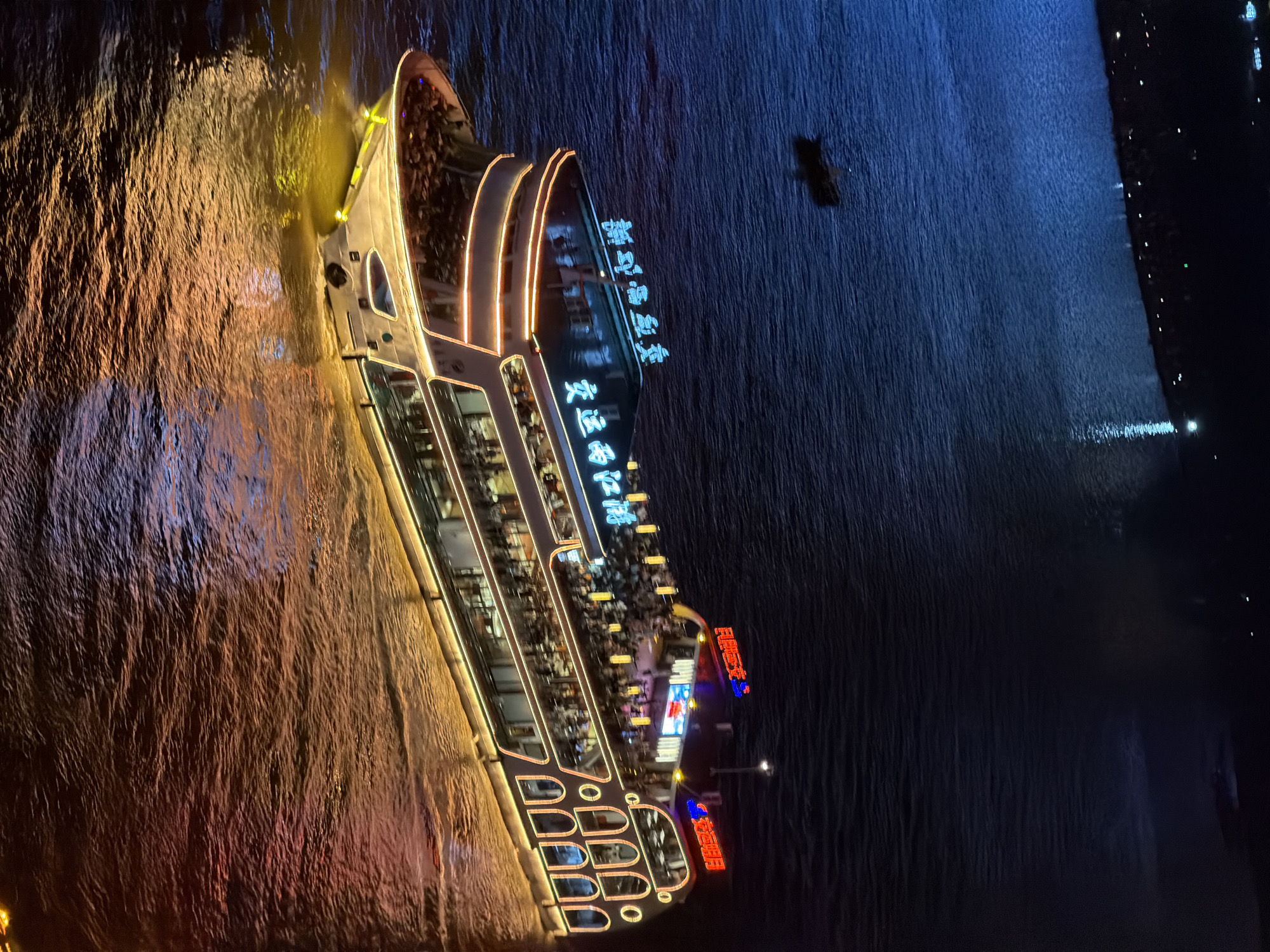 An aerial night view captures a brightly illuminated, multi-decked passenger ship navigating a dark body of water. The ship, with its white exterior, is adorned with extensive yellow and blue neon lighting, outlining its structure and windows. The upper decks are crowded with visible figures, suggesting passengers enjoying an evening outing. Chinese characters in neon, "新世纪明珠" and "交运两江", are displayed prominently on the ship's facade. The water's surface reflects the ship's lights, creating a warm orange and yellow glow. In the distance, a much smaller dark boat is visible on the left, and faint lights suggest a shoreline on the upper left of the frame. The overall scene depicts a vibrant urban nightscape on a river, likely within the Yuzhong District of Chongqing, China, a location known for its dramatic riverfront scenery and extensive nighttime illumination.